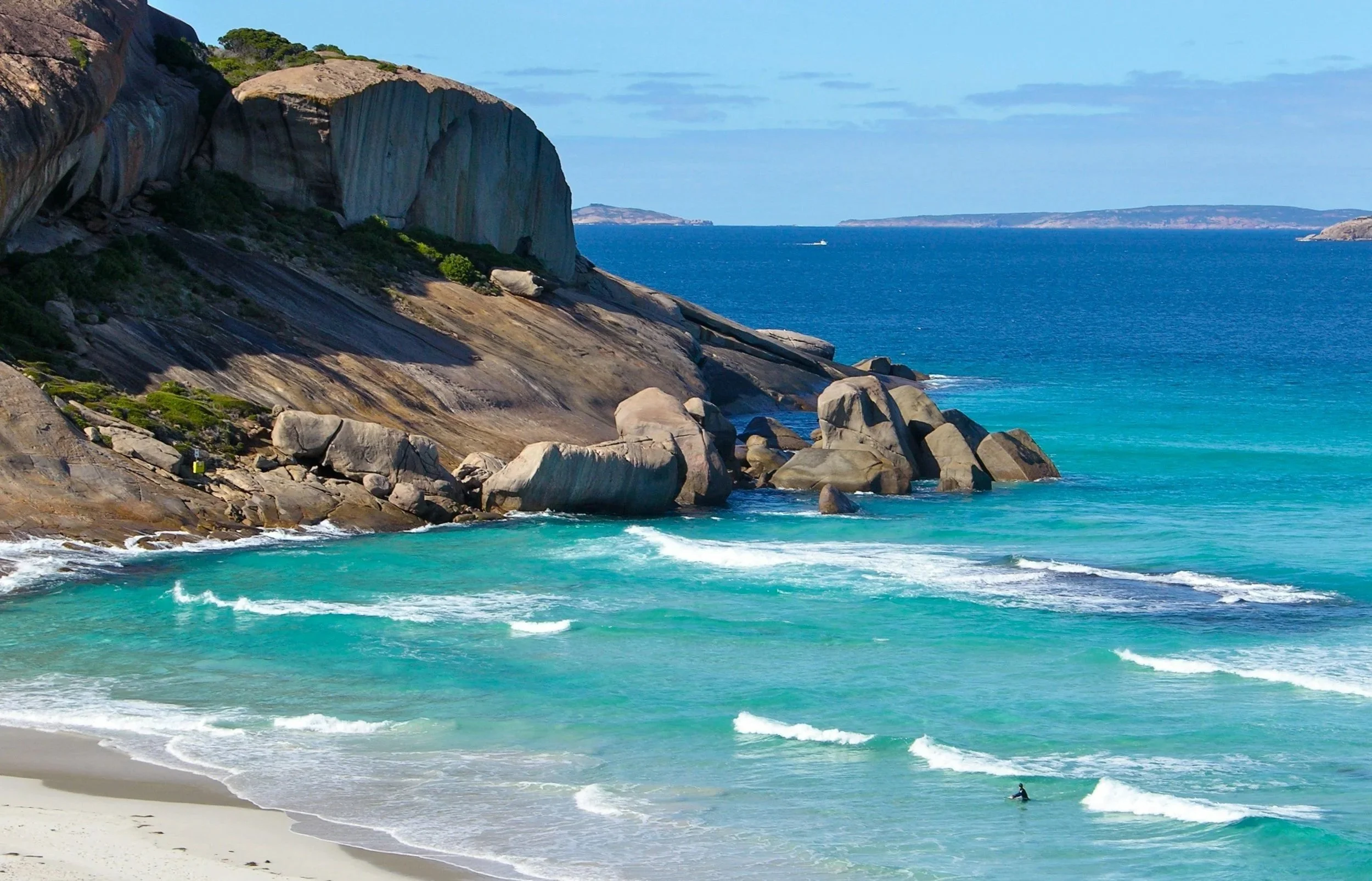A scenic view of a beach with turquoise waters, large rock formations along the shoreline, and cliffs in the background under a partly cloudy sky.