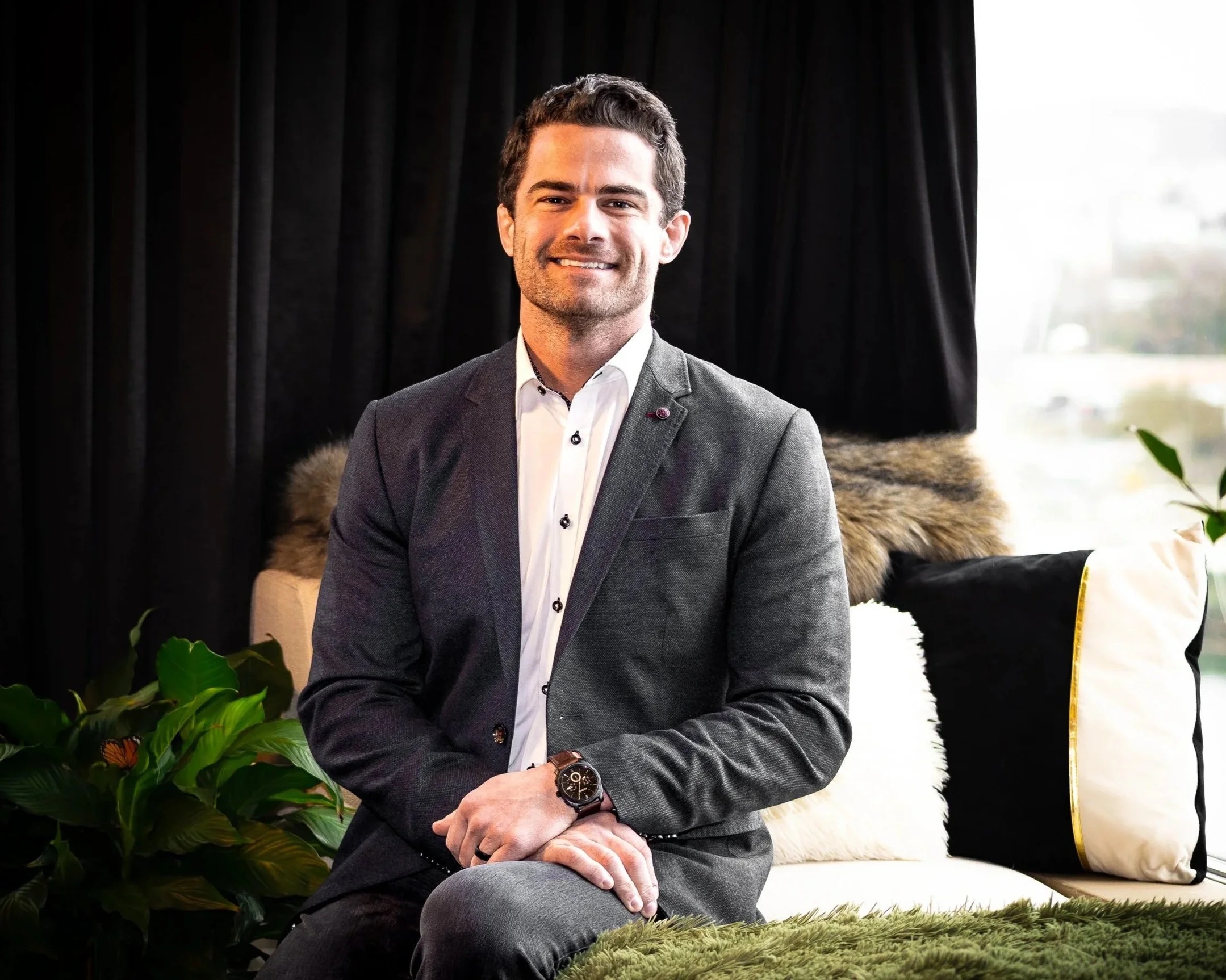 A man in a gray suit and white shirt sits on a couch with black, white, and fur pillows, next to a green plant, in a room with black curtains and a window with daylight.