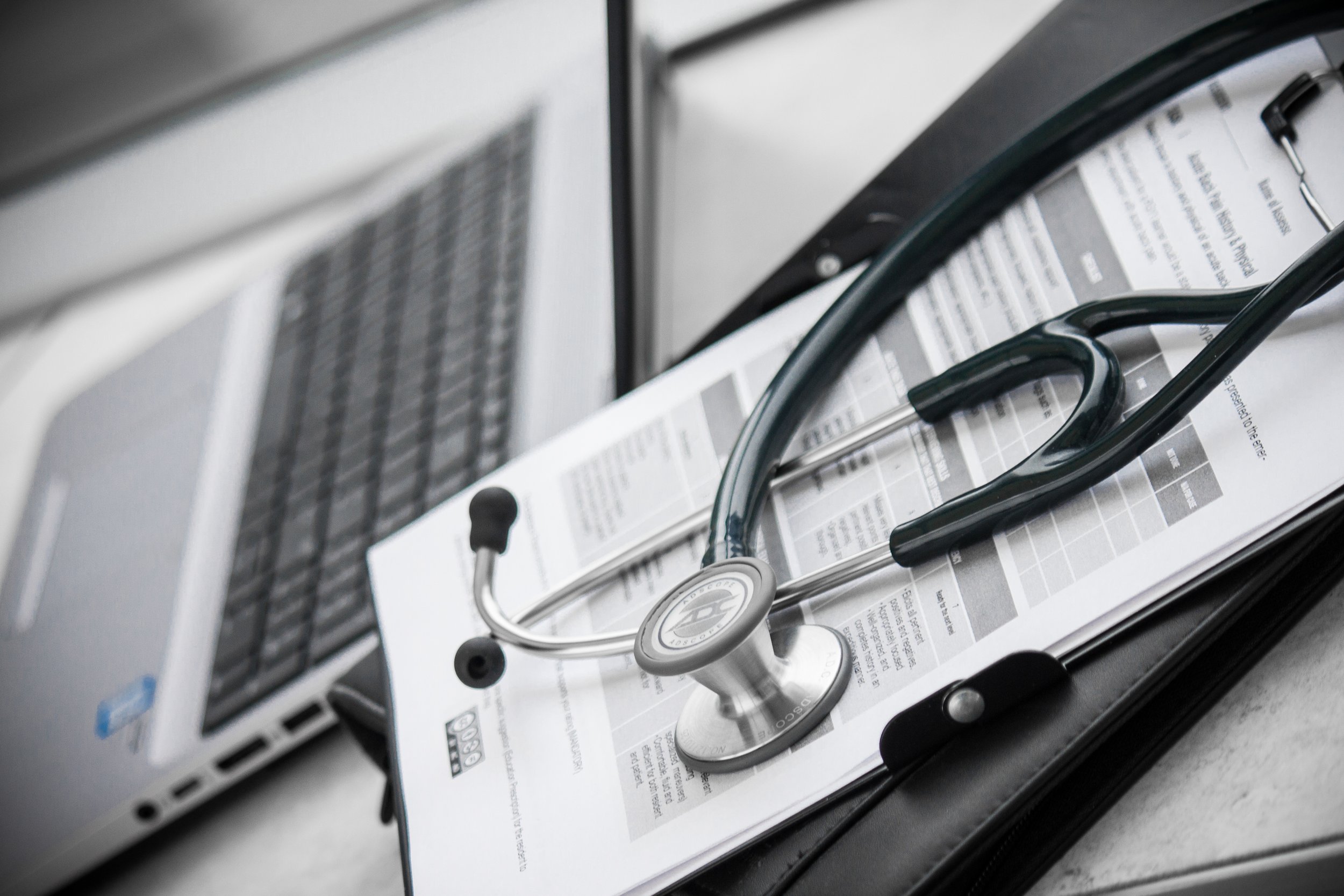 A stethoscope resting on a clipboard with medical documents, placed on a gray surface near a laptop.