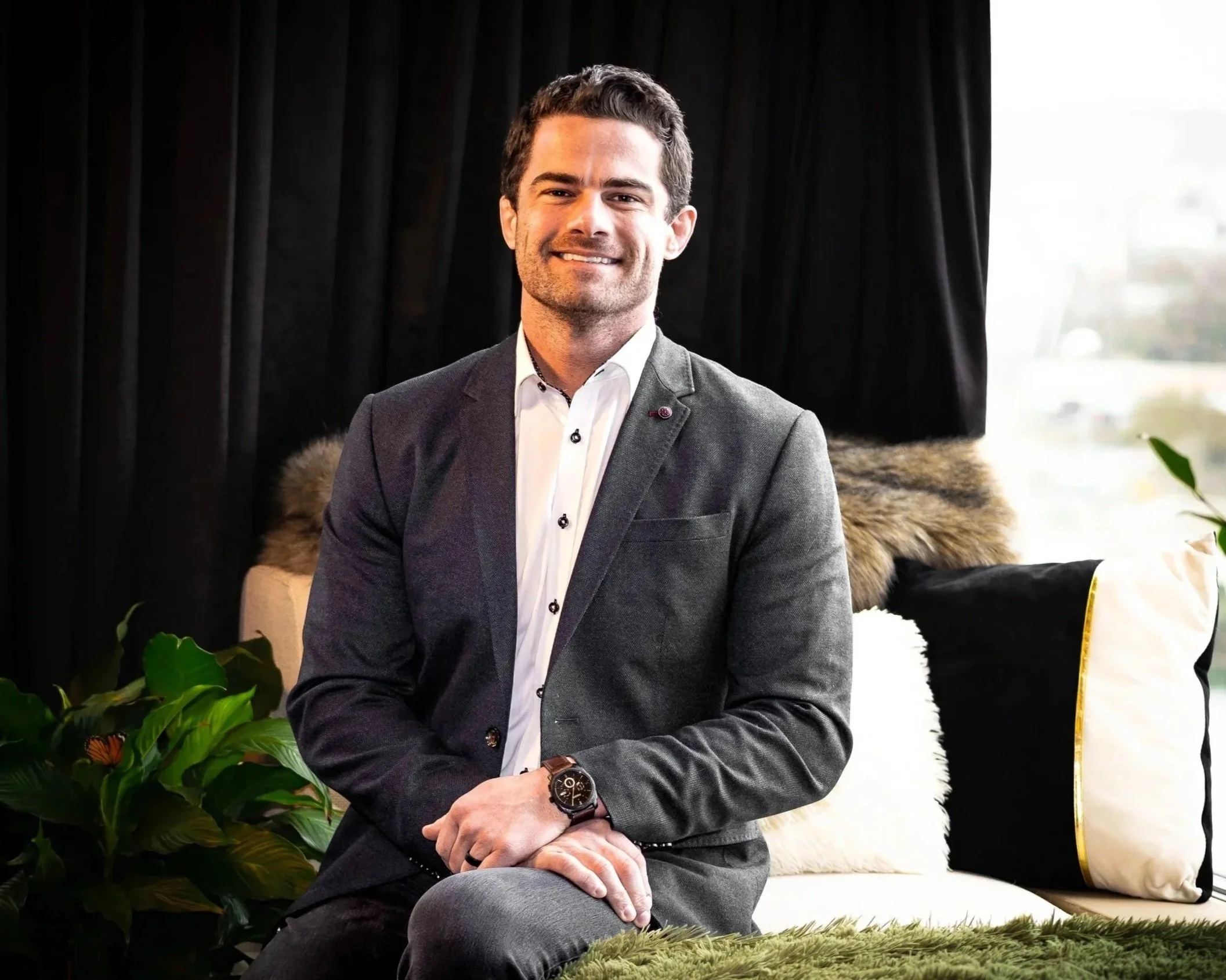 A man in a gray suit with a white shirt sitting on a green plush cushion, smiling in a modern room with black curtains, plants, and pillows including black, white, and a furry one.