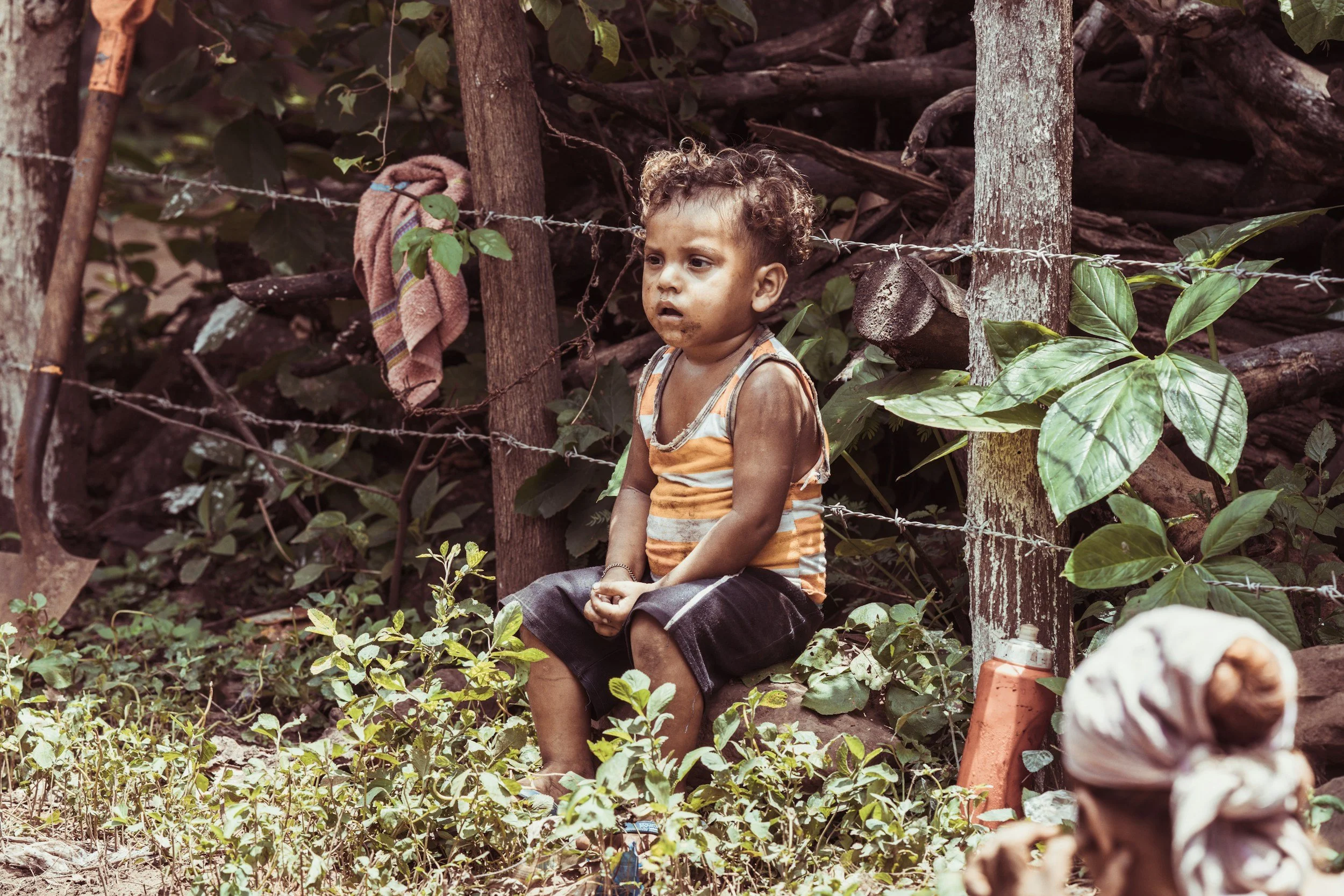 A young boy with curly hair sits on the ground in a wooded area, looking distressed, with dirty face and hands, wearing a sleeveless striped shirt and dark shorts, behind a barbed wire fence with wooden posts, surrounded by green plants and trees.