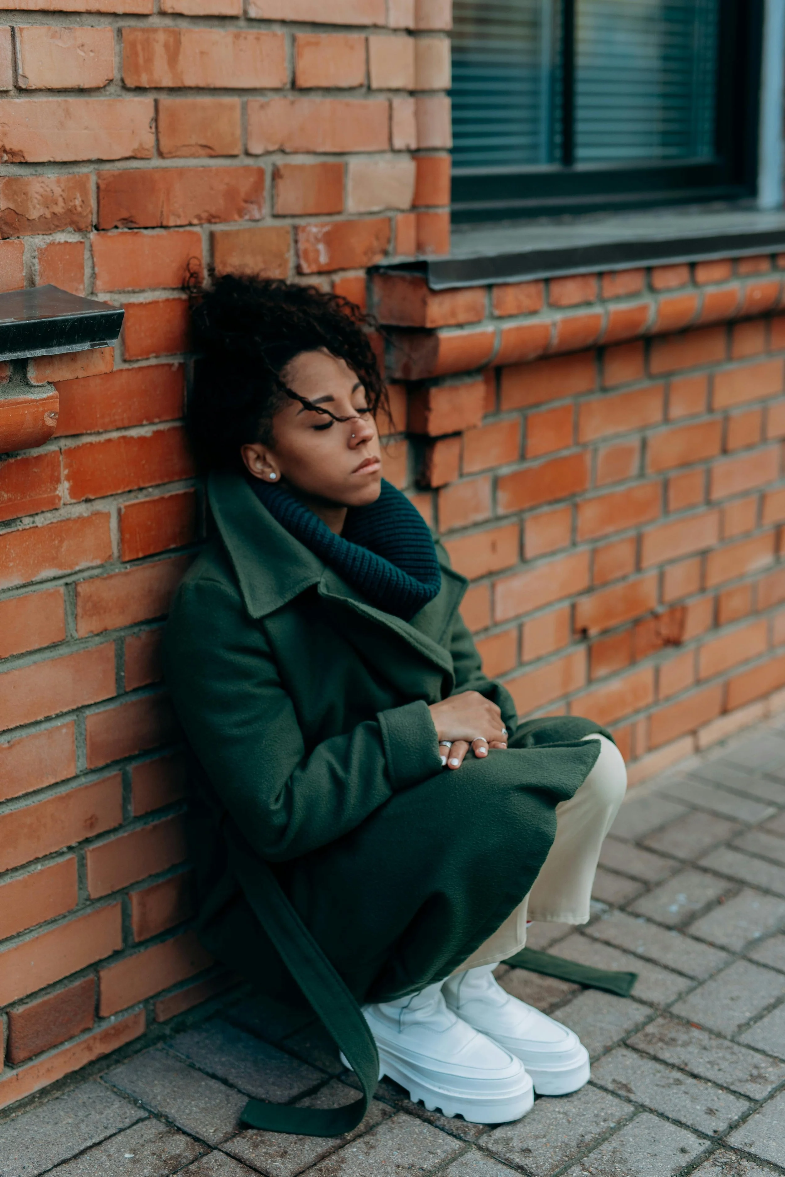 A woman with curly hair, sitting on the sidewalk, leaning against a brick wall, her eyes closed, wearing a green coat, black scarf, beige pants, and white sneakers.