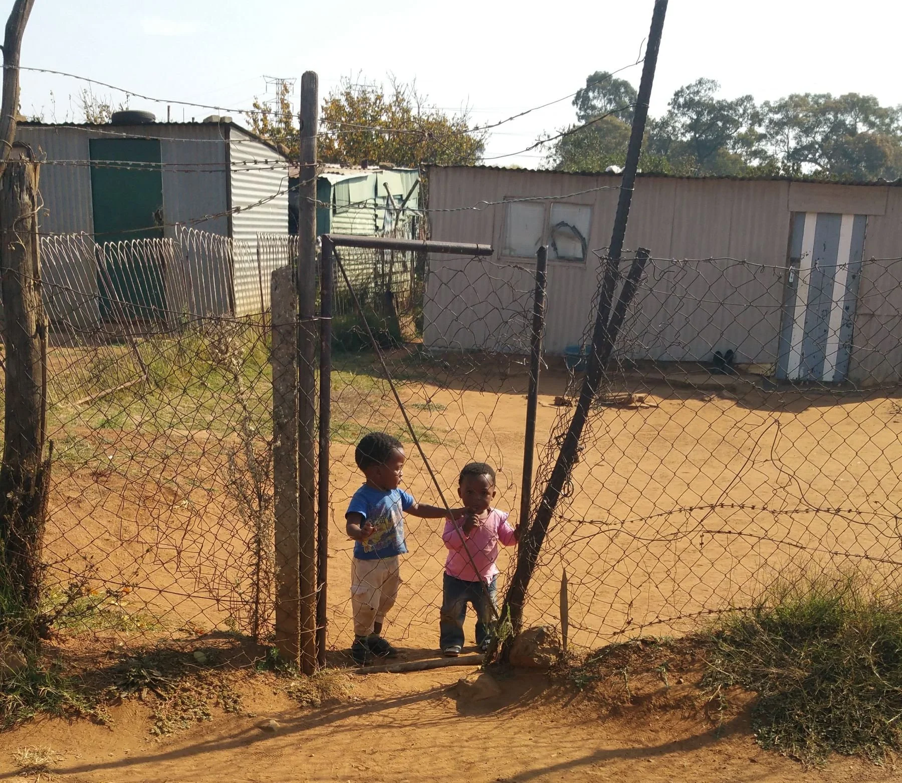 Two young children standing behind a makeshift fence made of wire, with one child touching the other's chest. The background features simple buildings and dirt ground.