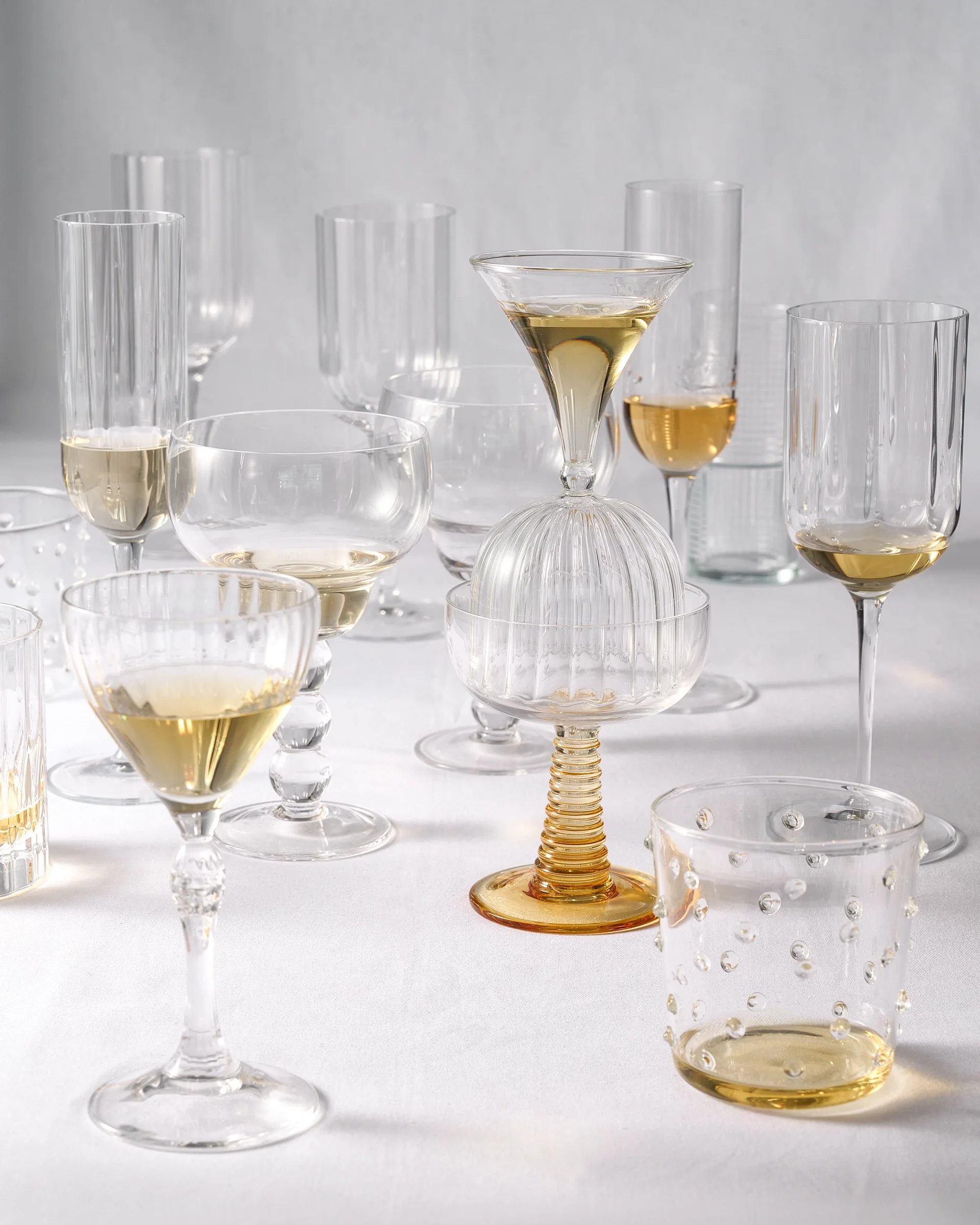 a collection of varied, elegant glassware on a white tablecloth, including ribbed coupes, wine glasses, and a unique amber-stemmed glass