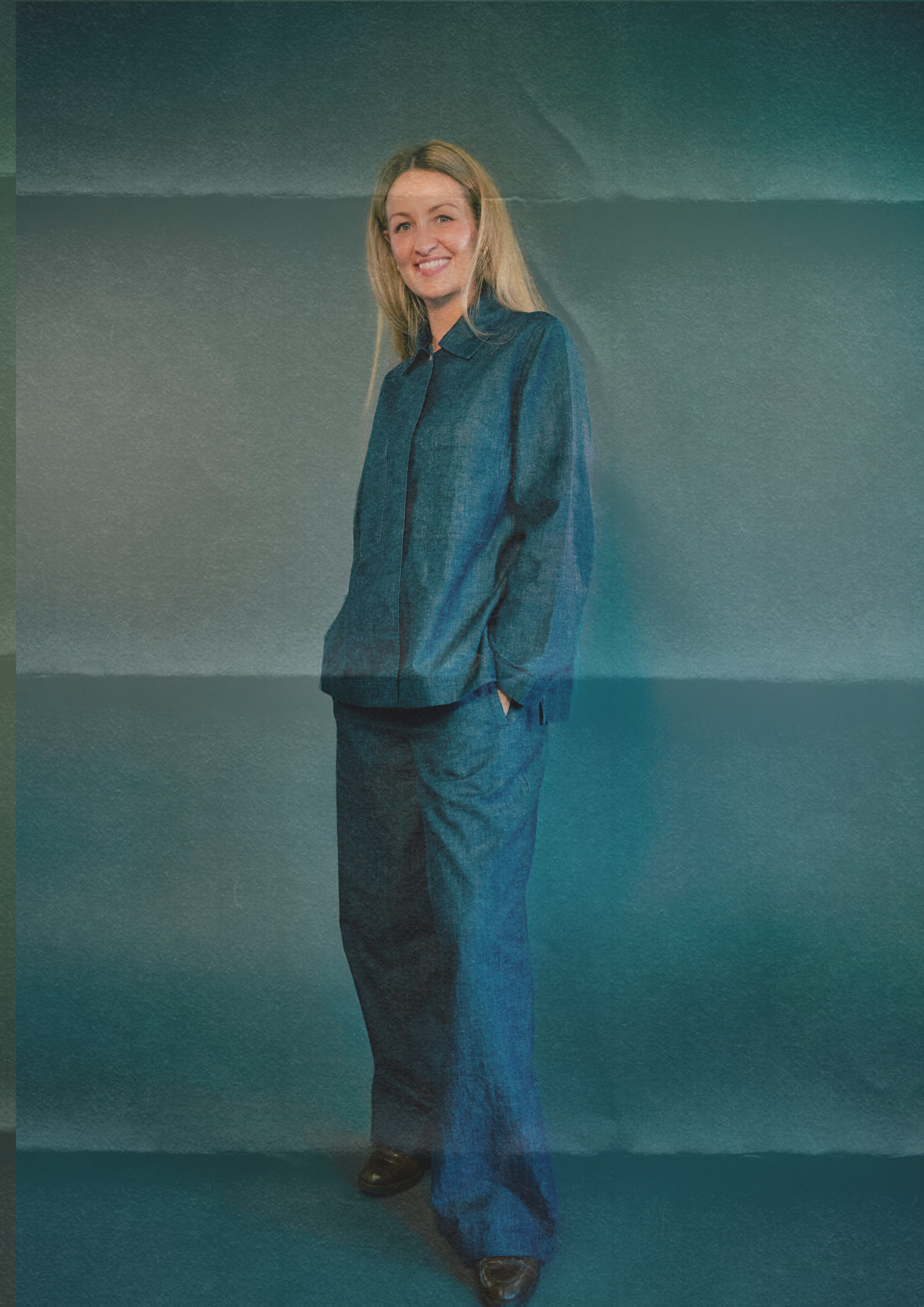 Eine lächelnde Frau in blauer Hose und Blazer steht vor einer grauen Wand.