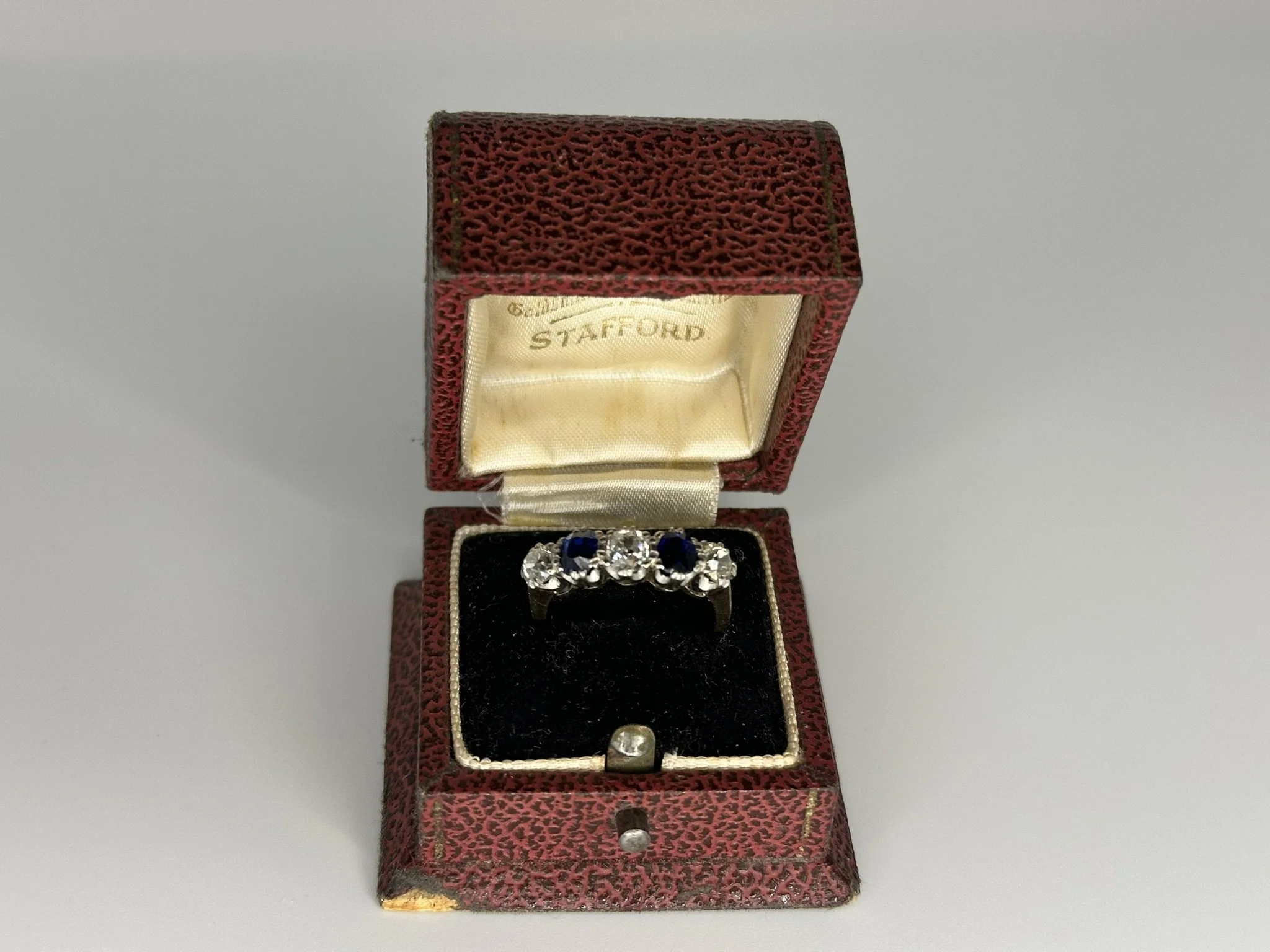 A vintage red jewelry box with black interior contains a silver ring with four gemstones, two sapphires and two diamonds, placed on a soft black cushion. The inside of the lid is lined with cream satin fabric with gold lettering that reads "STAFFORD." The box has a textured exterior and shows signs of wear on the corners.