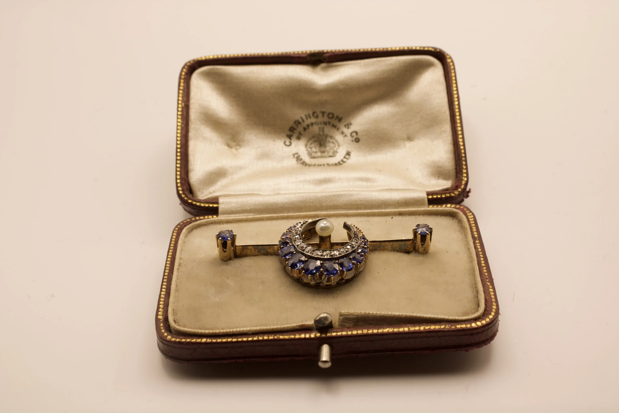 Vintage jewelry box with an ornate brooch inside, decorated with blue and clear gemstones and a pearl, on a white surface.
