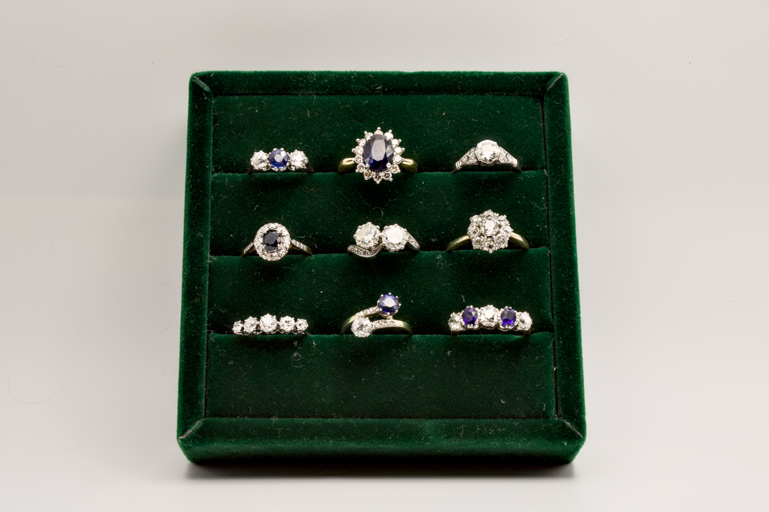 A green ring display box filled with nine vintage-style rings featuring diamonds and sapphires.