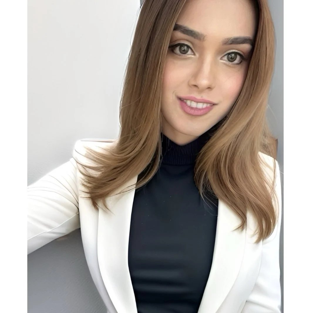 A young woman with light brown hair and gray eyes taking a selfie indoors, wearing a black turtleneck and a white blazer.