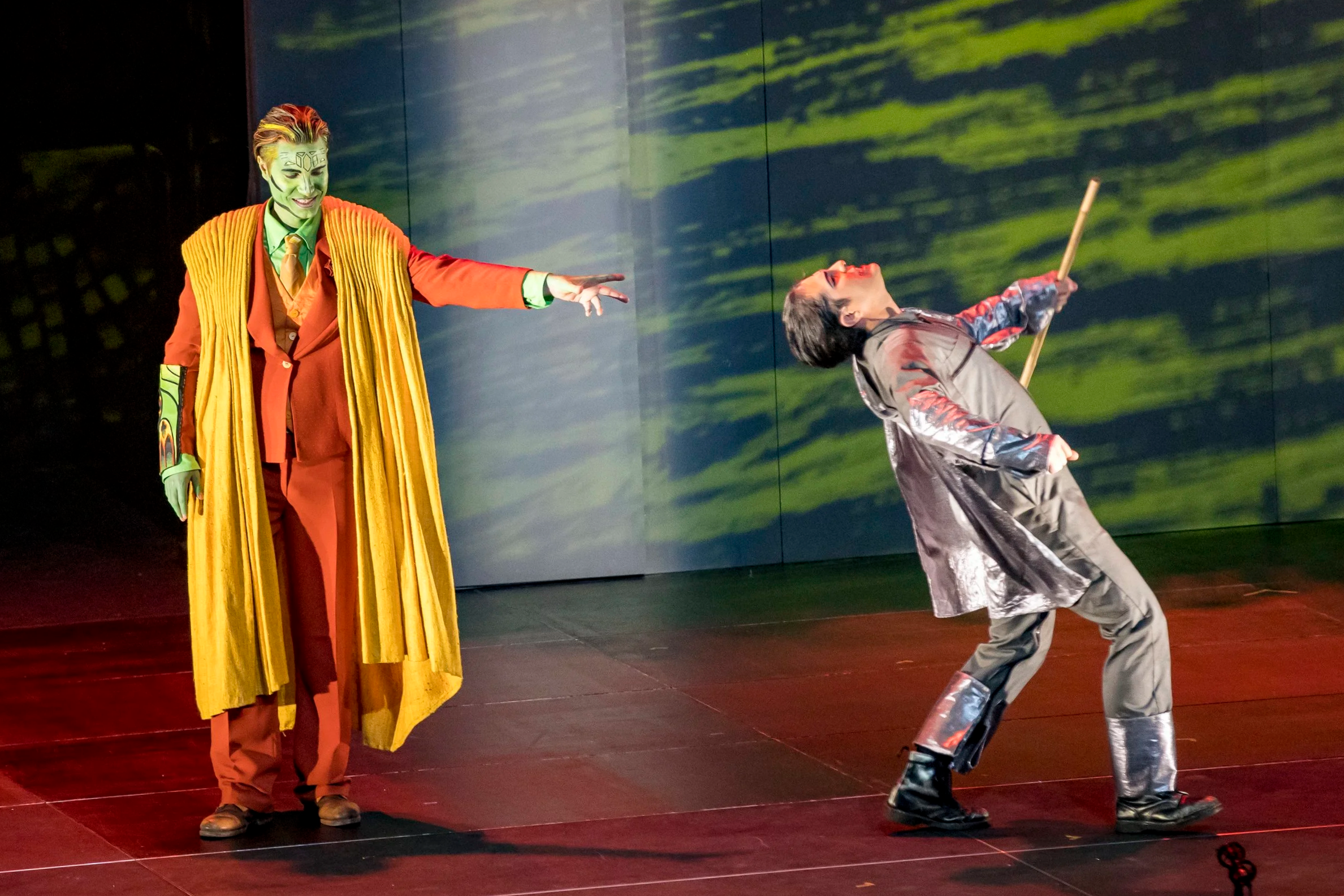 Deux acteurs en costume coloré sont en scène, l'un avec un maquillage vert et un manteau jaune, pointant vers une autre personne vêtue d'une veste grise et tenant un bâton, qui est incliné en arrière.