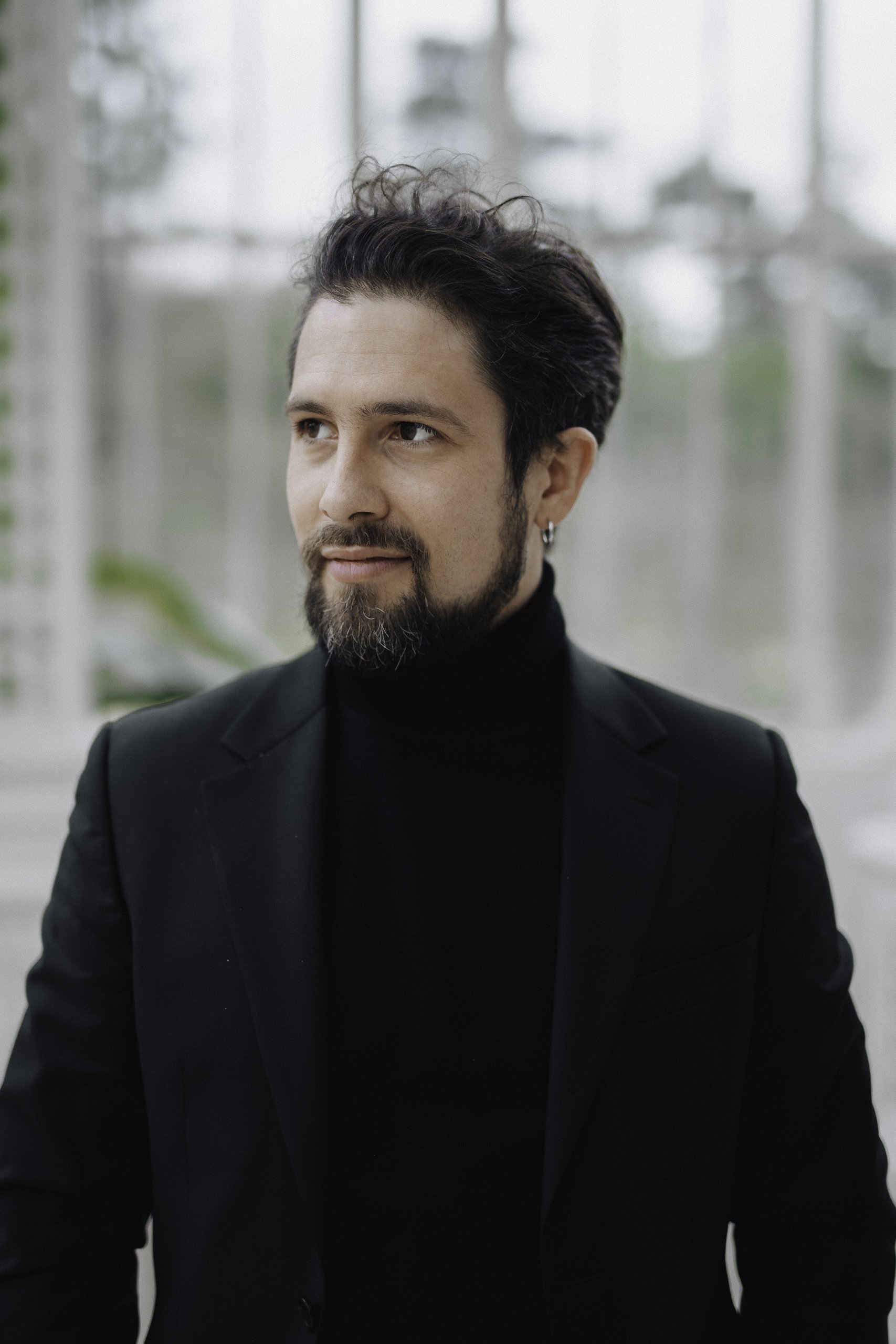 Un homme avec une barbe, portant un costume noir et un col roulé noir, assis devant une grande fenêtre avec un arrière-plan flou de nature.
