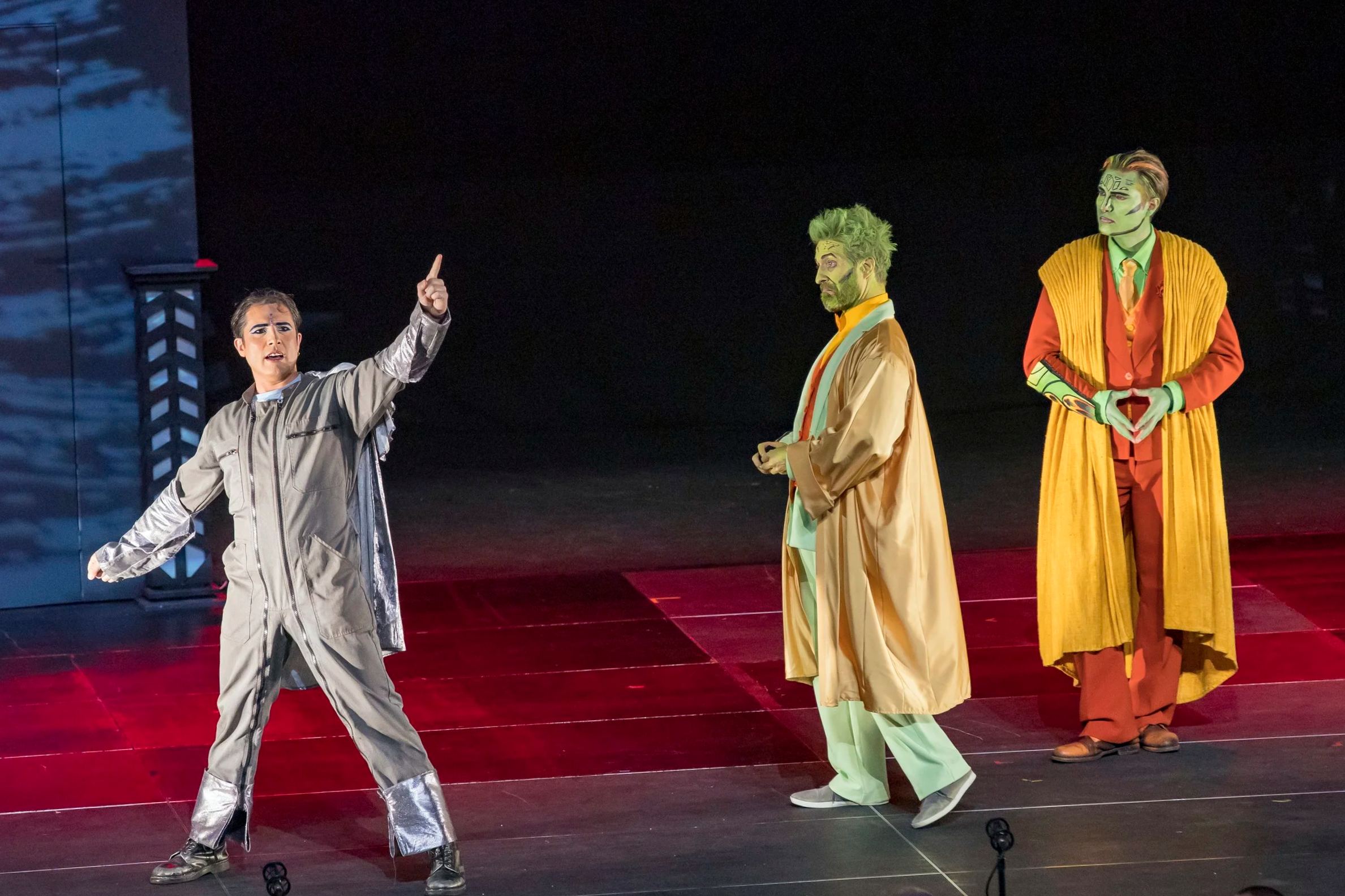 Scène de théâtre avec trois acteurs en costumes colorés sur scène, trois hommes, en pleine représentation.