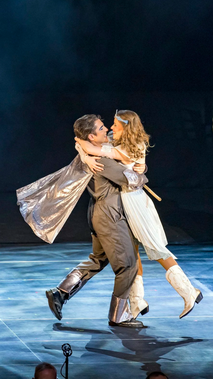 Deux acteurs sur scène, un homme en costume gris et une femme en costume blanc, se tiennent dans une étreinte joyeuse, dans le contexte d'une performance théâtrale, avec un fond noir et un éclairage de scène.