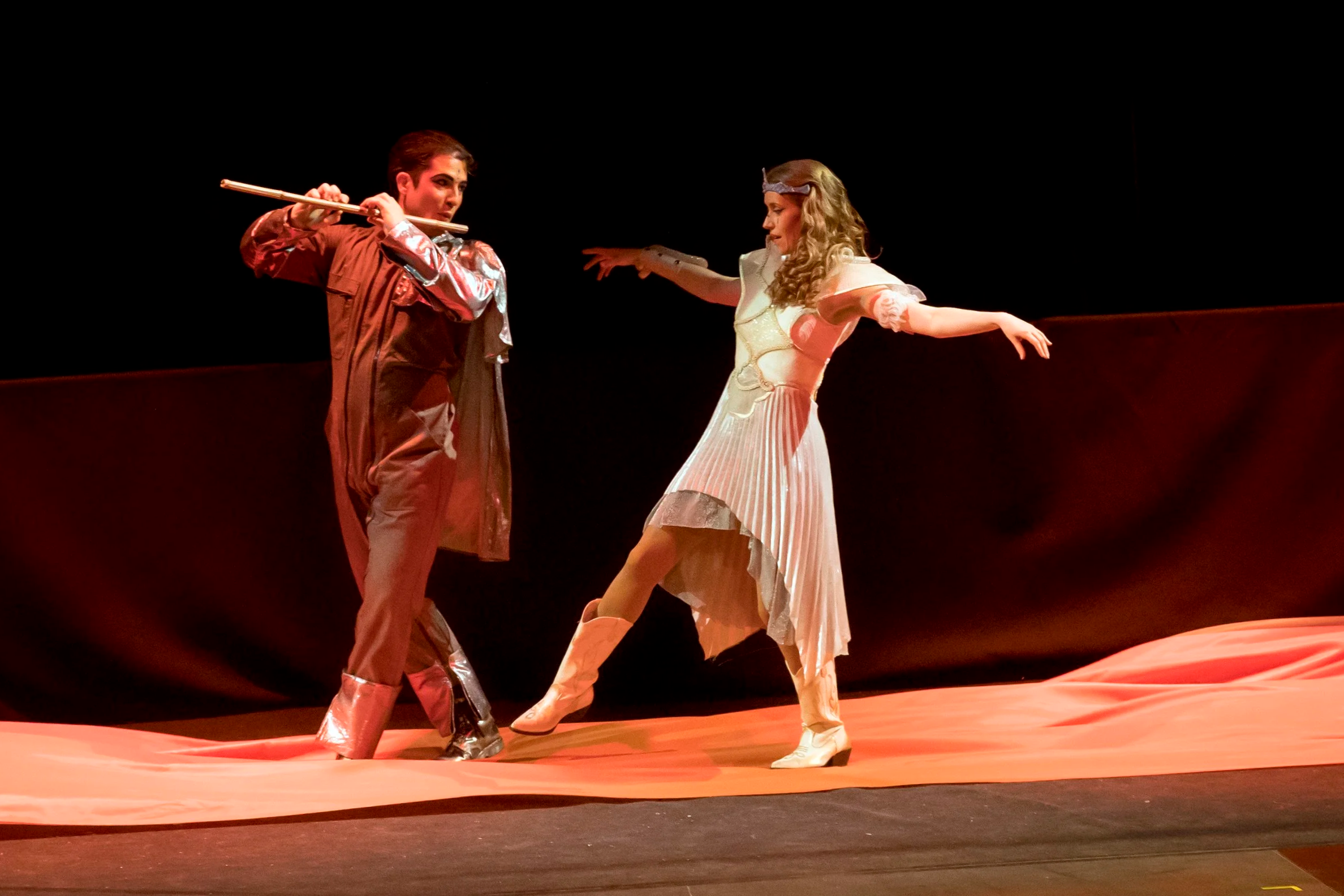 Tamino et Pamina, dans La Flûte Enchantée de Mozart, en costumes de théâtre dramatiques. Tamino joue d'une flûte en roseau, en pleine scène de théâtre sur un fond noir et un sol en tissu orange.