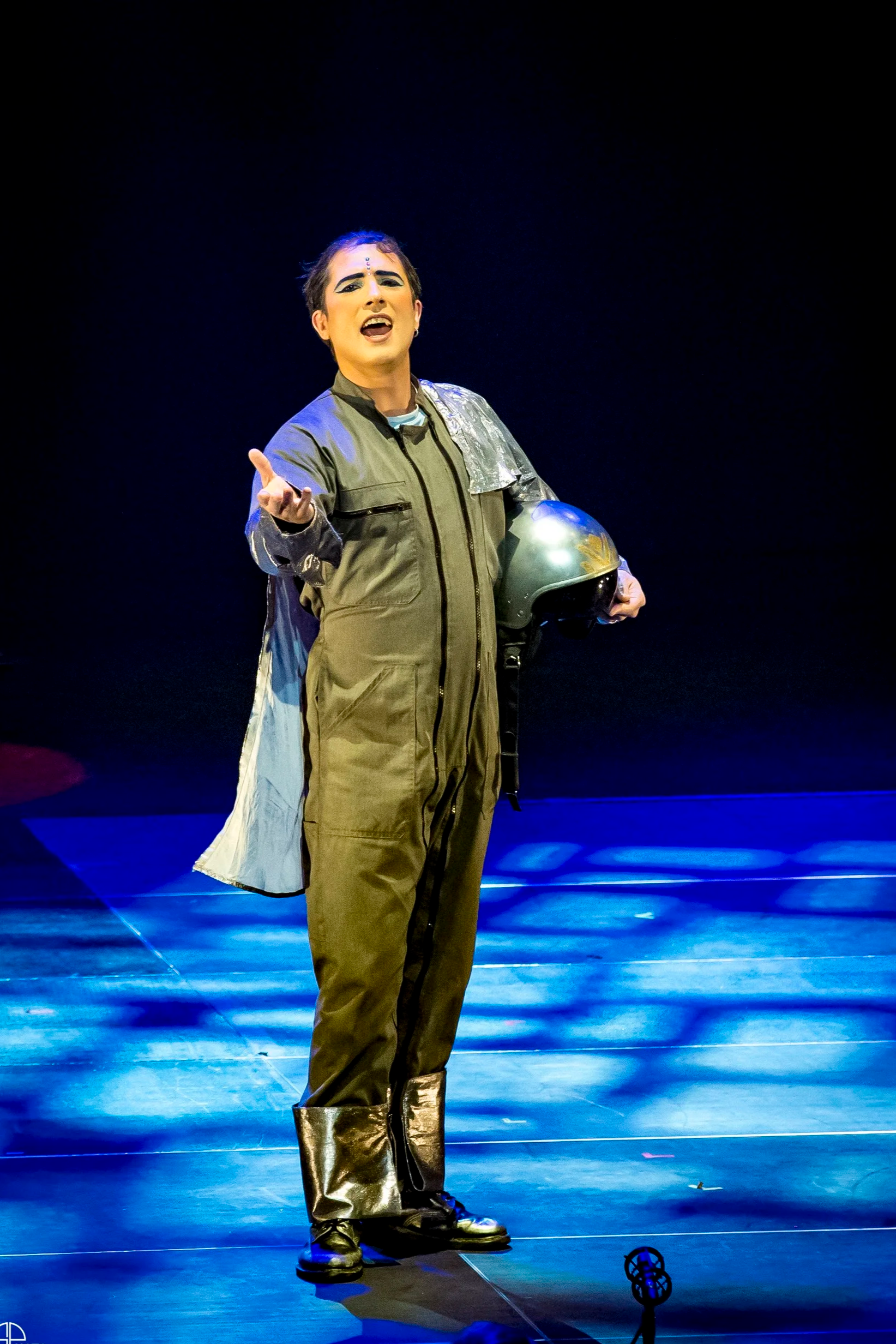 Une personne en costume représentant un cosmonaute super-héros, tenant un casque dans une main et étendant l'autre vers l'avant, dans une représentation de La Flûte Enchantée de Mozart avec un éclairage coloré.
