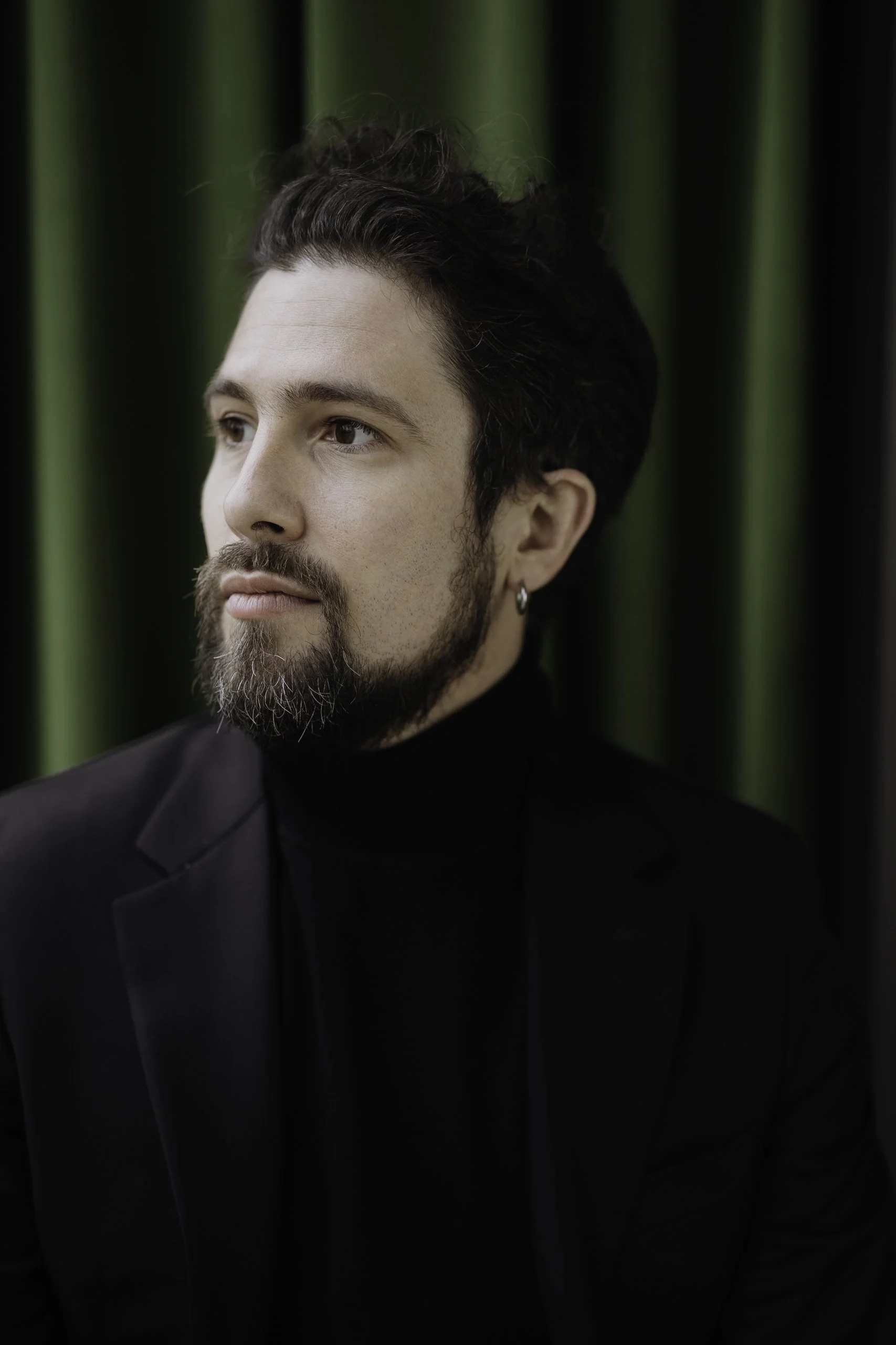 Un homme avec une barbe, des cheveux foncés et bouclés, portant un blazer noir et un col roulé noir, pose devant un rideau vert foncé.