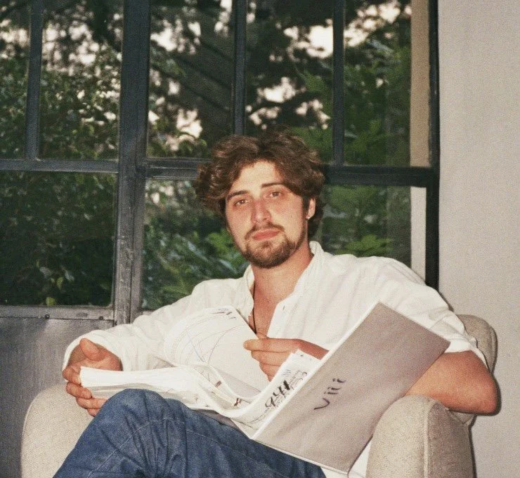 A young man with wavy brown hair, a beard, and wearing a white shirt, sitting in a beige armchair near a large window with black framing. He is holding an open newspaper or magazine with some visible text and looking at the camera.