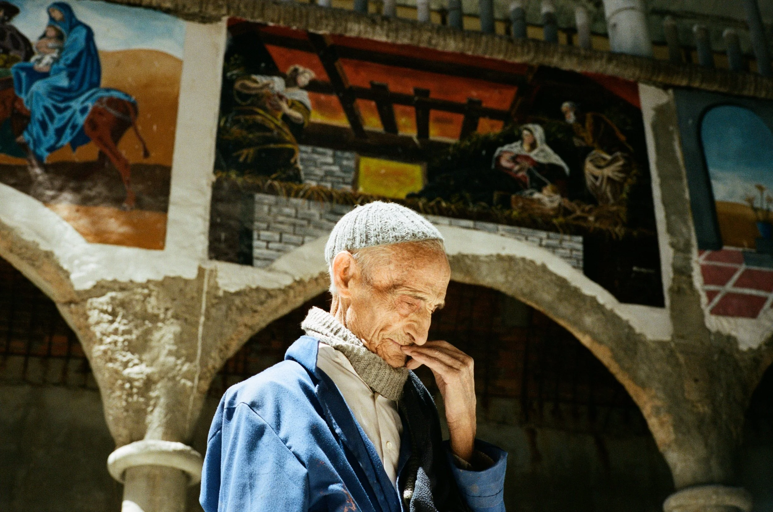 An elderly man with a gray yarmulke and blue jacket, smiling and touching his face, standing in front of a wall with religious murals.