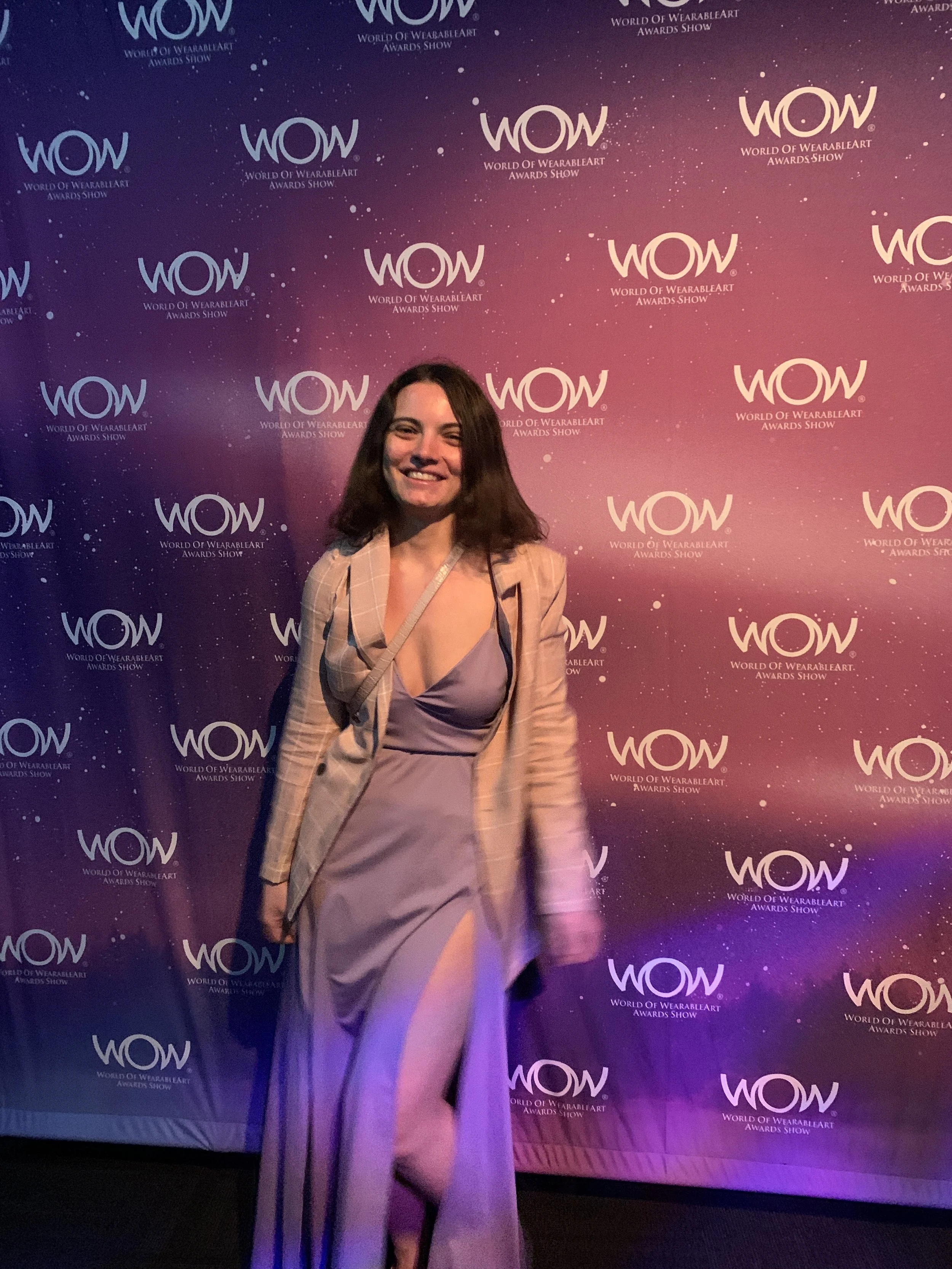 A woman smiling in front of a purple and pink gradient backdrop with the WOW logo and text 'World of WearableArt Awards Show.'