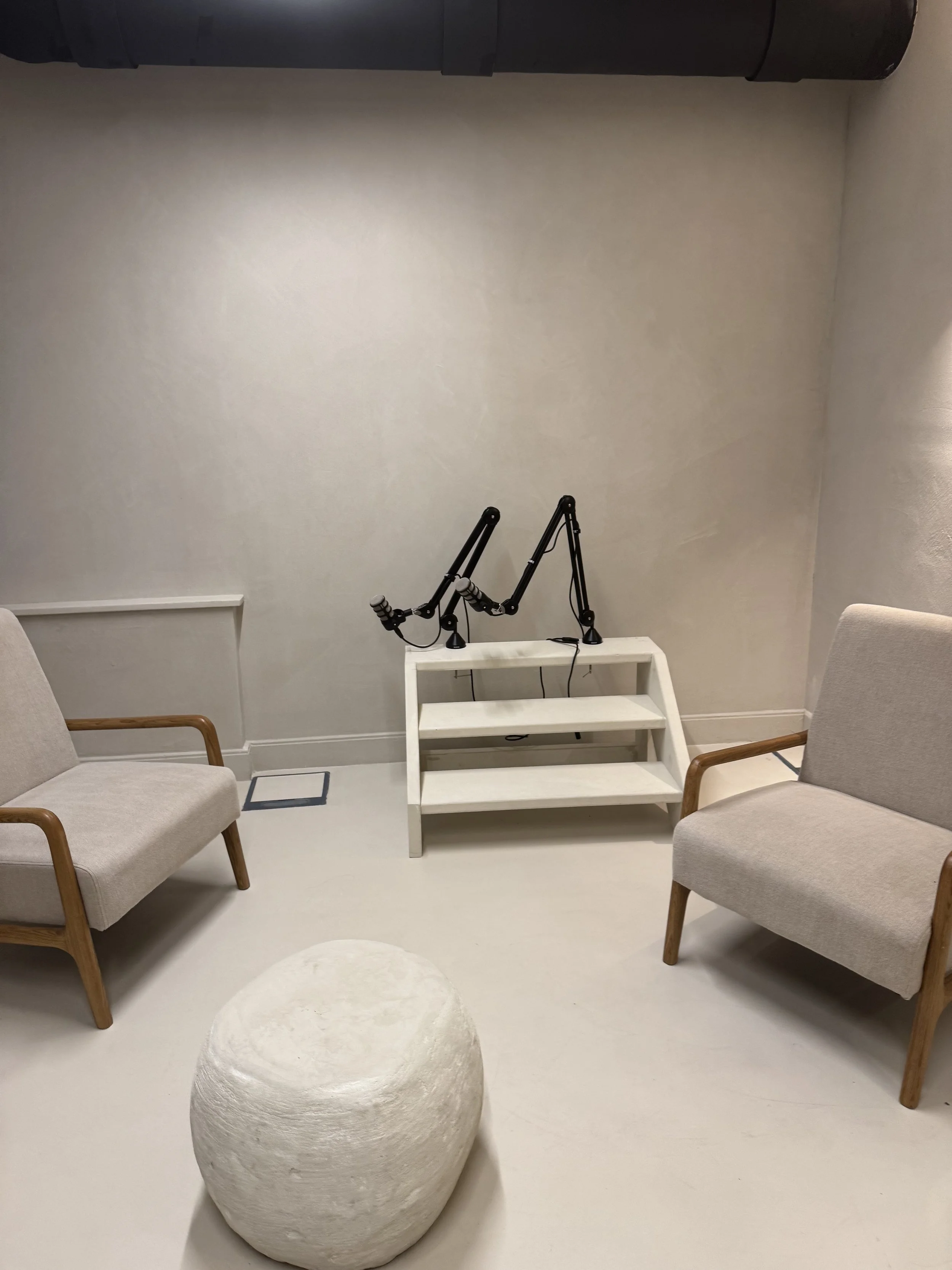 A minimalistic room with two beige armchairs with wooden arms, a white stool, a white shelving unit, and a microphone arm stand on top of the shelf. The room has white walls and flooring, with a dark pipe along the ceiling.