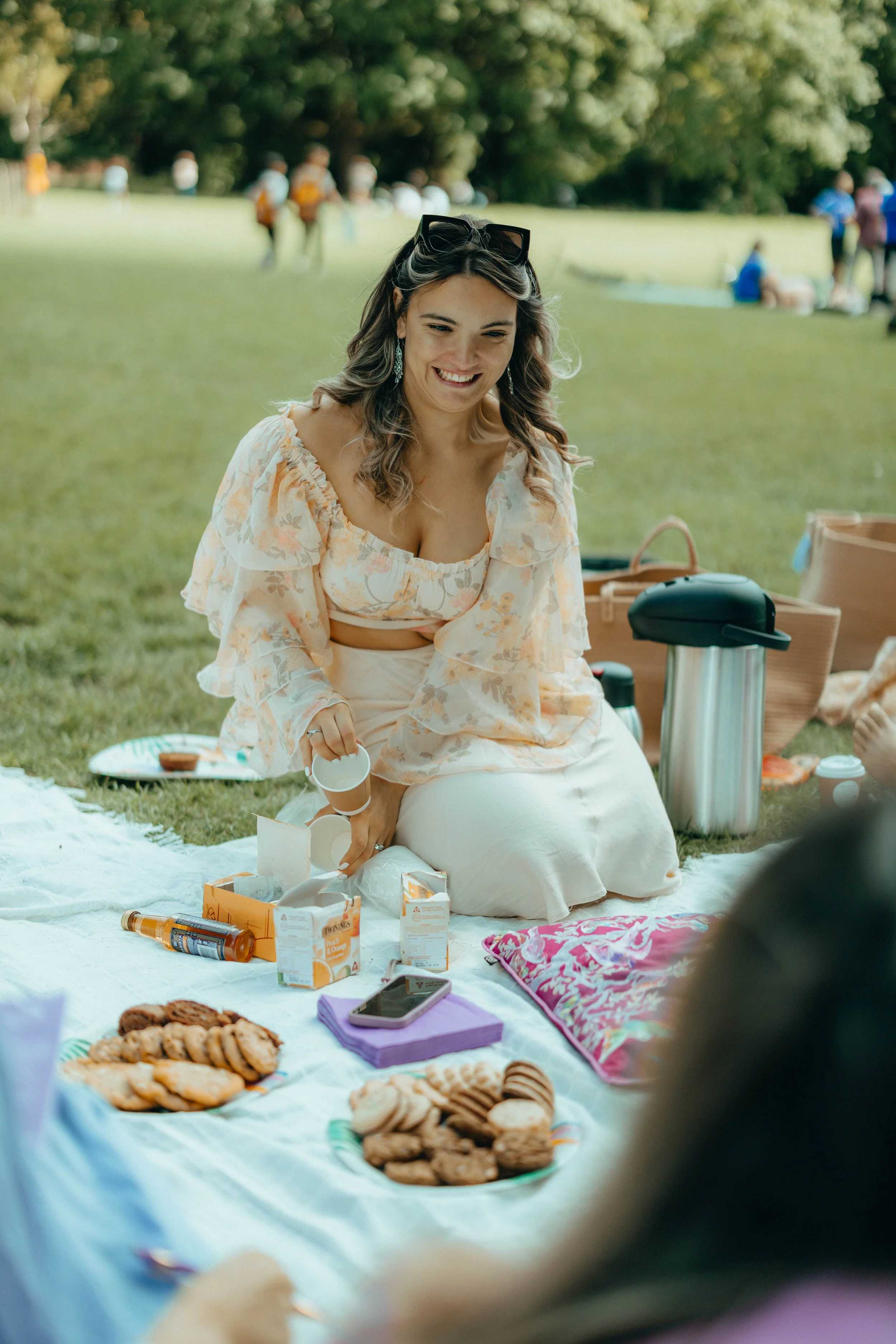 A woman sitting on a picnic blanket at a park, smiling, with plates of cookies, drinks, and a tea set around her.
