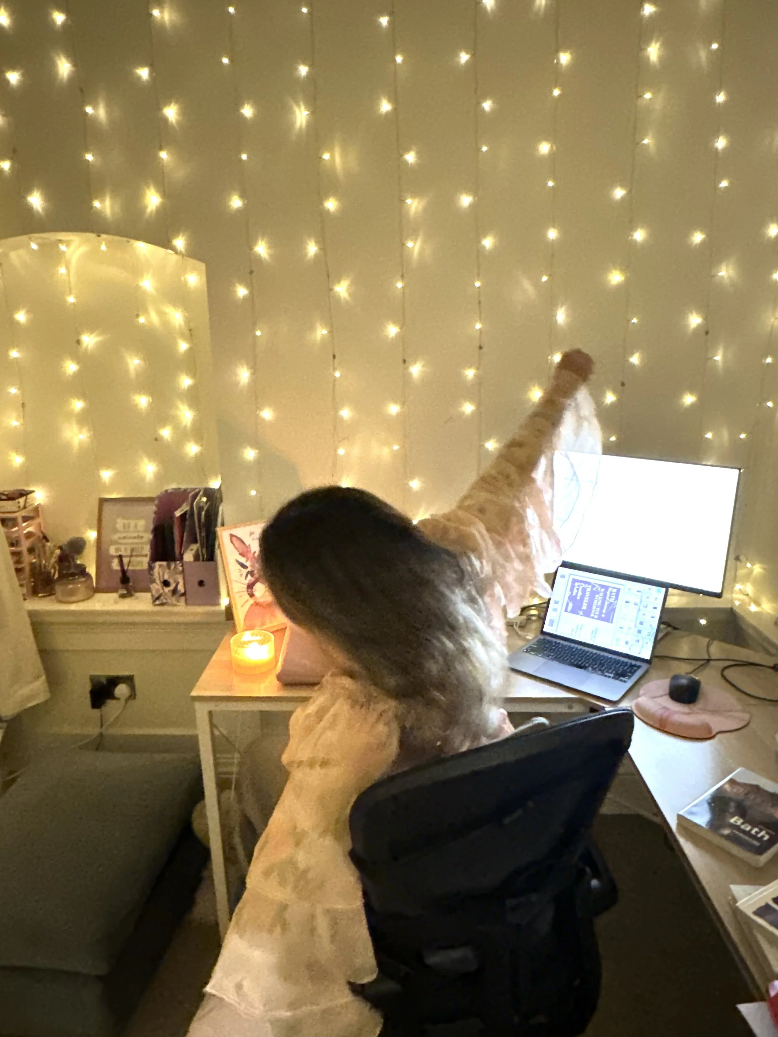 A person with dark curly hair sitting at a desk in front of two screens, one large and one smaller, with a fairy light wall in the background. The desk has a candle, books, and other items.