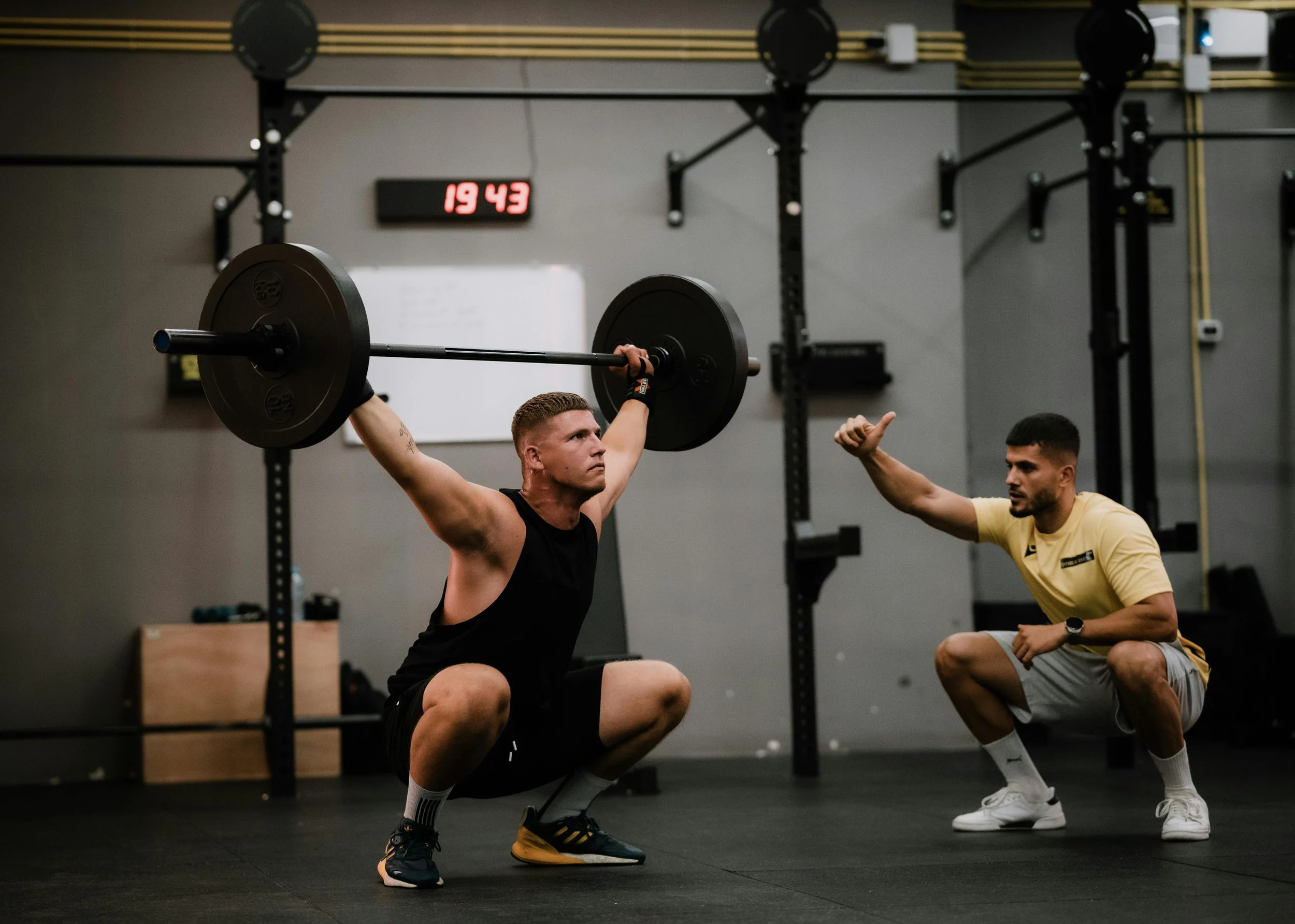 Man in black tank top performing a back squat with a barbell in a gym, coach nearby giving instructions