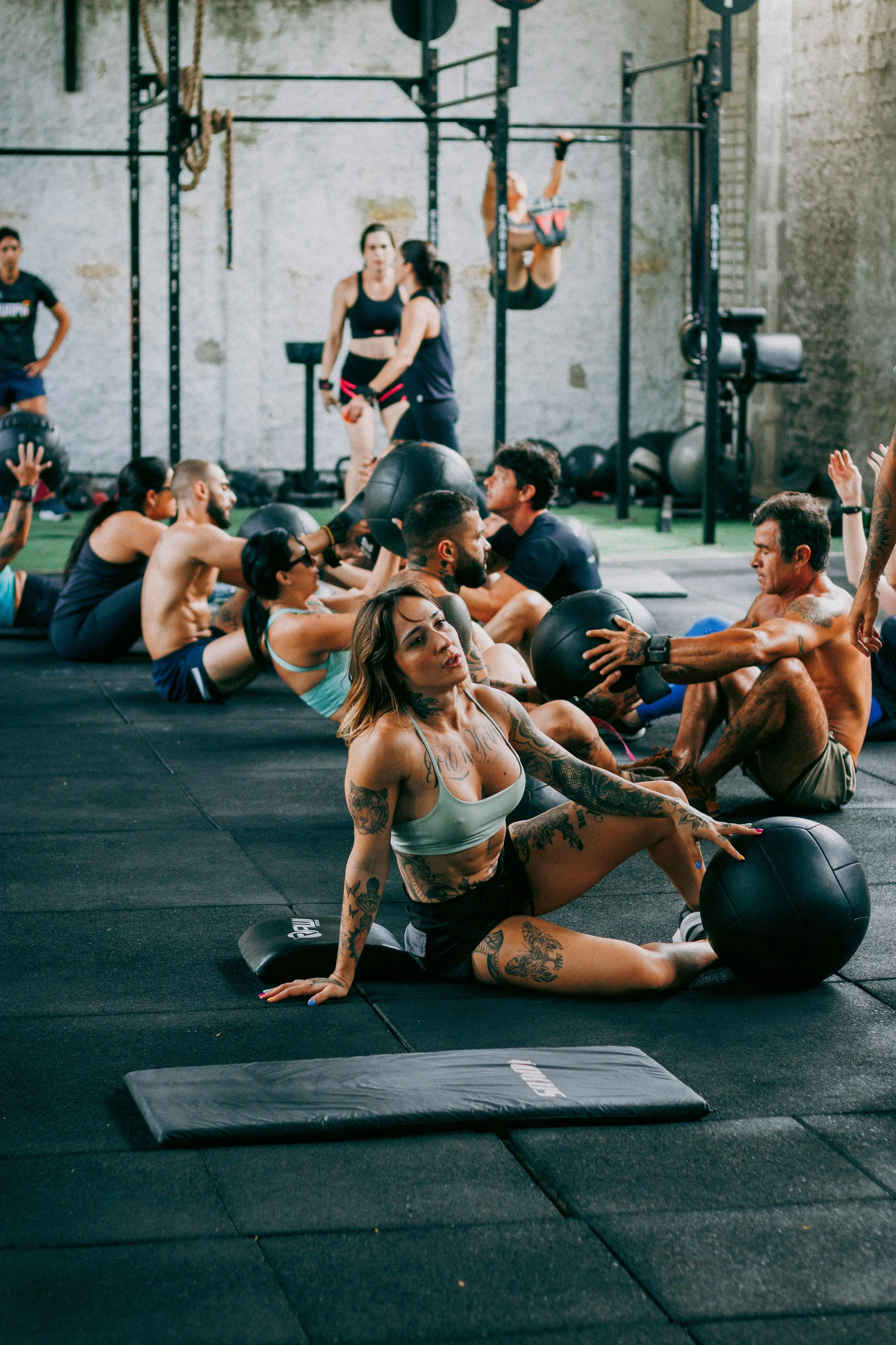 People working out in a gym, doing sit-ups with medicine balls on a black mat, with others lifting weights in the background.