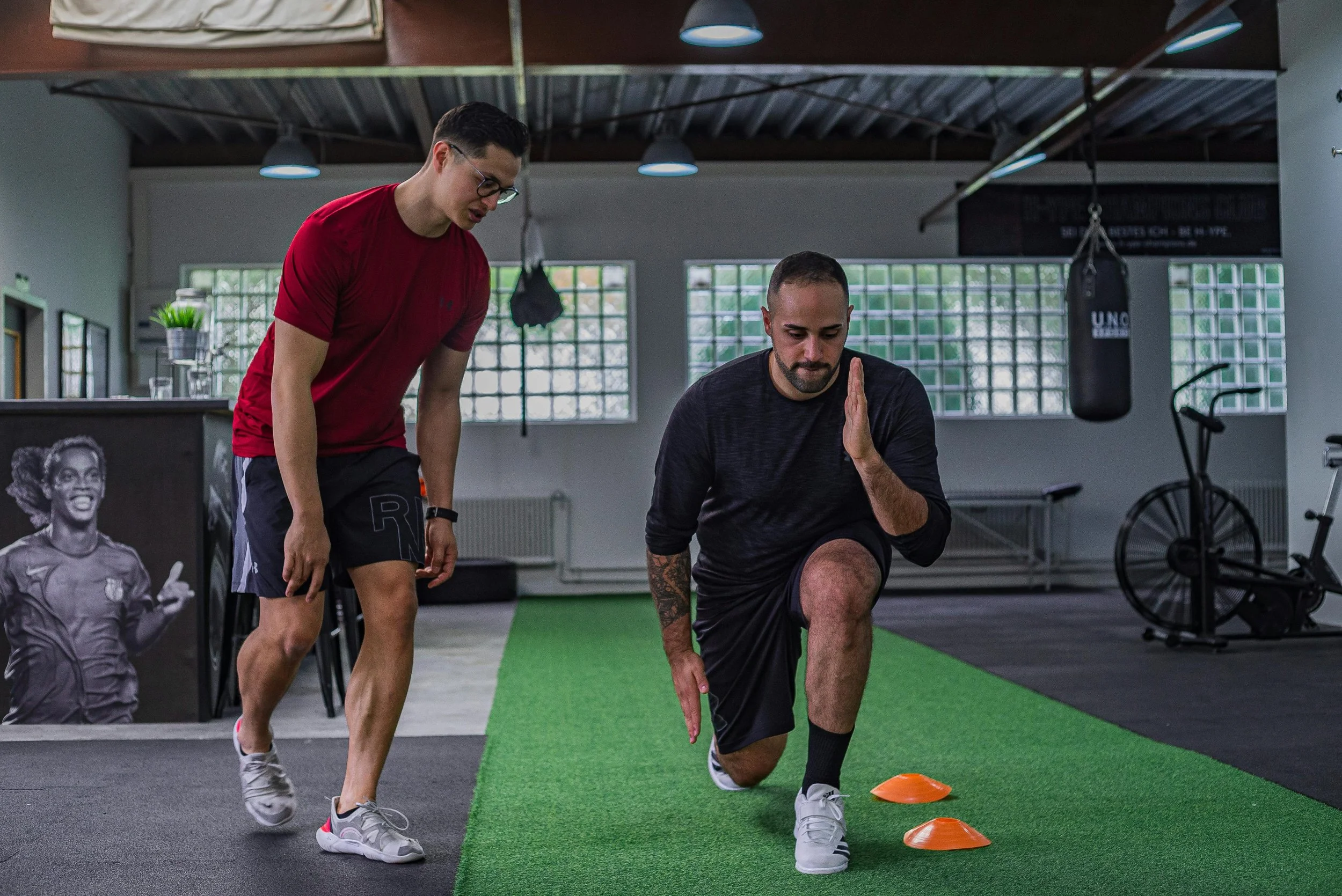 A trainer assisting a man in a gym with a cone drill exercise on a green turf. The man is on one knee, balancing on one leg, with a hand raised in front of him. The gym has large windows, punching bag, and exercise equipment in the background.