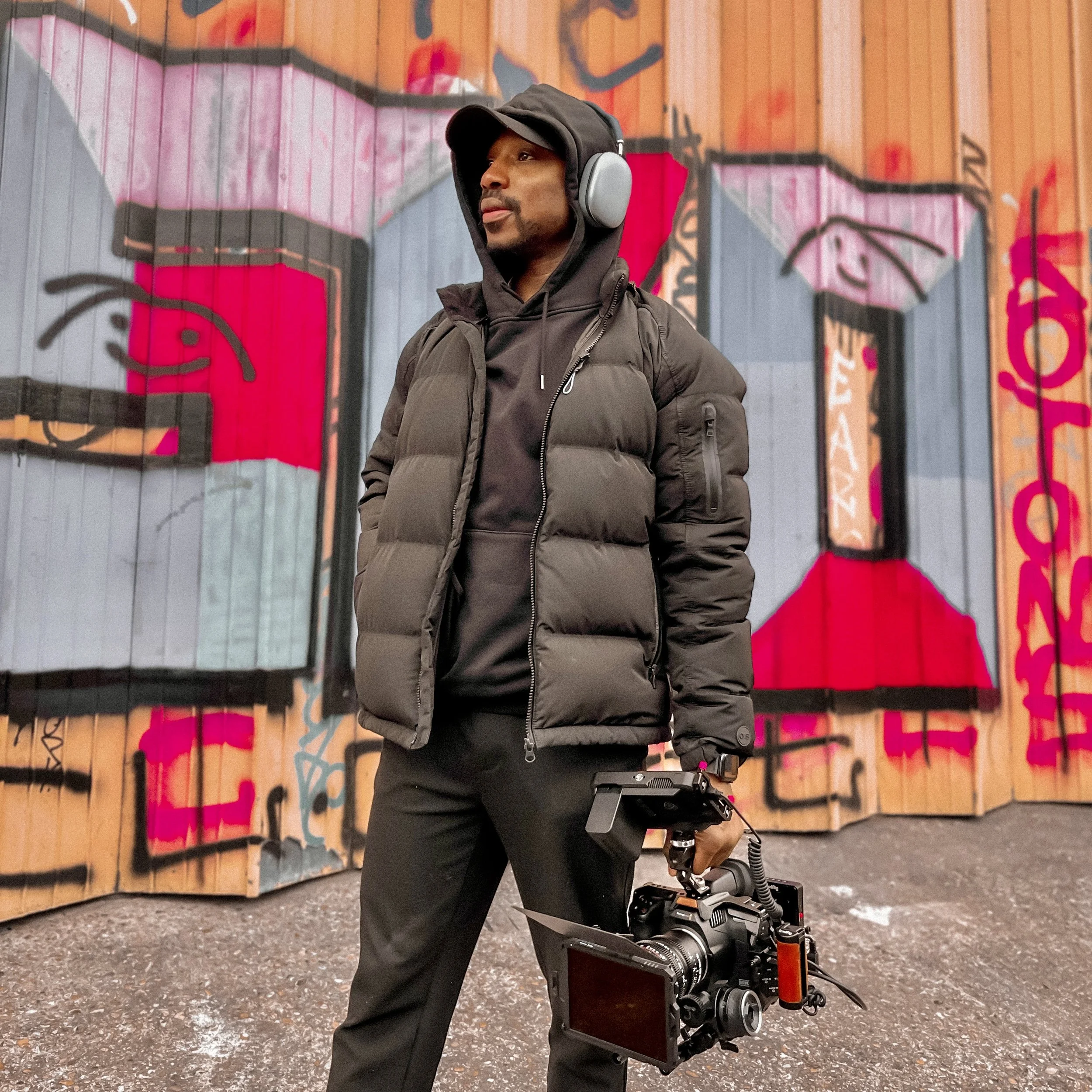 A man in black hoodie, puffer jacket, and headphones holding a professional video camera in front of a colorful graffiti wall.