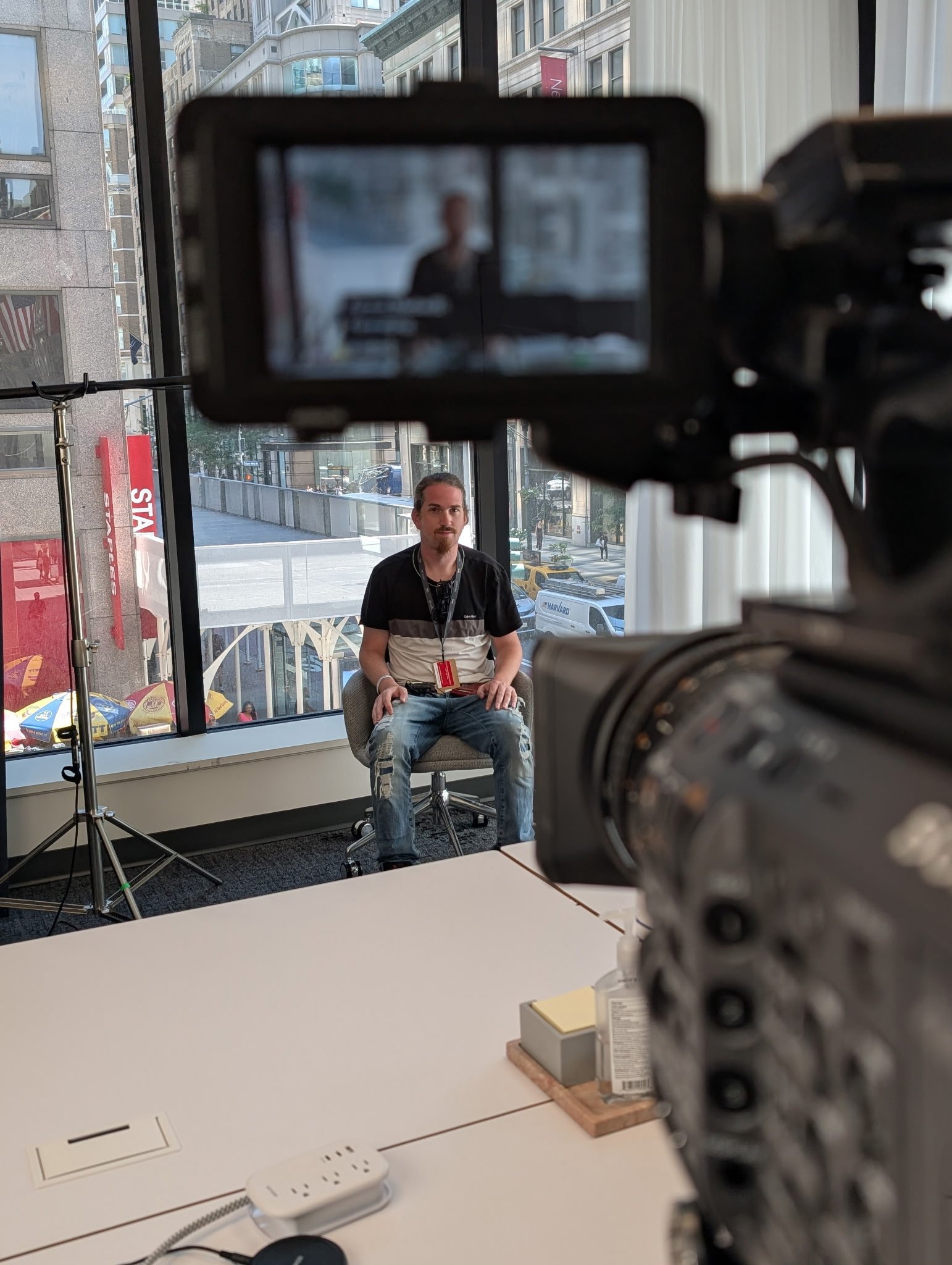 A man sitting on a chair in front of a large window, with a city street outside, is being filmed or photographed with a professional camera in an indoor setting.