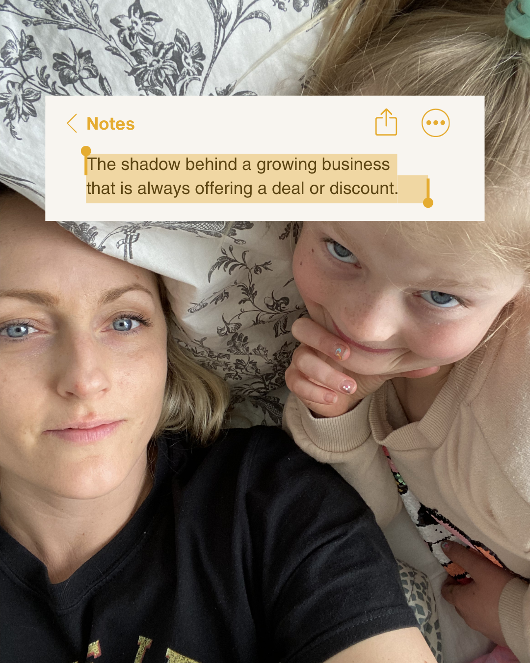 A woman with blonde hair and a young girl with blonde hair lying on a bed or couch, taking a selfie together. The woman is wearing a black shirt, and the girl is wearing a beige hoodie with colorful designs. The woman has clear skin and blue eyes, and the girl has blue eyes, freckles, and a small rainbow and pearl nail design on her fingers.