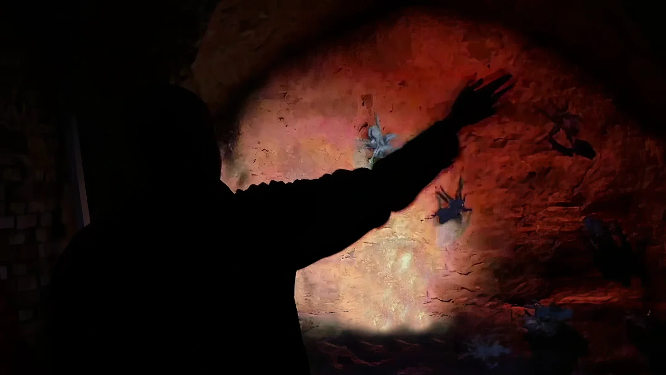 Silhouette of a person reaching out with their hand, touching a wall with black leaf illustrations, illuminated by reddish lighting in a dark indoor setting.