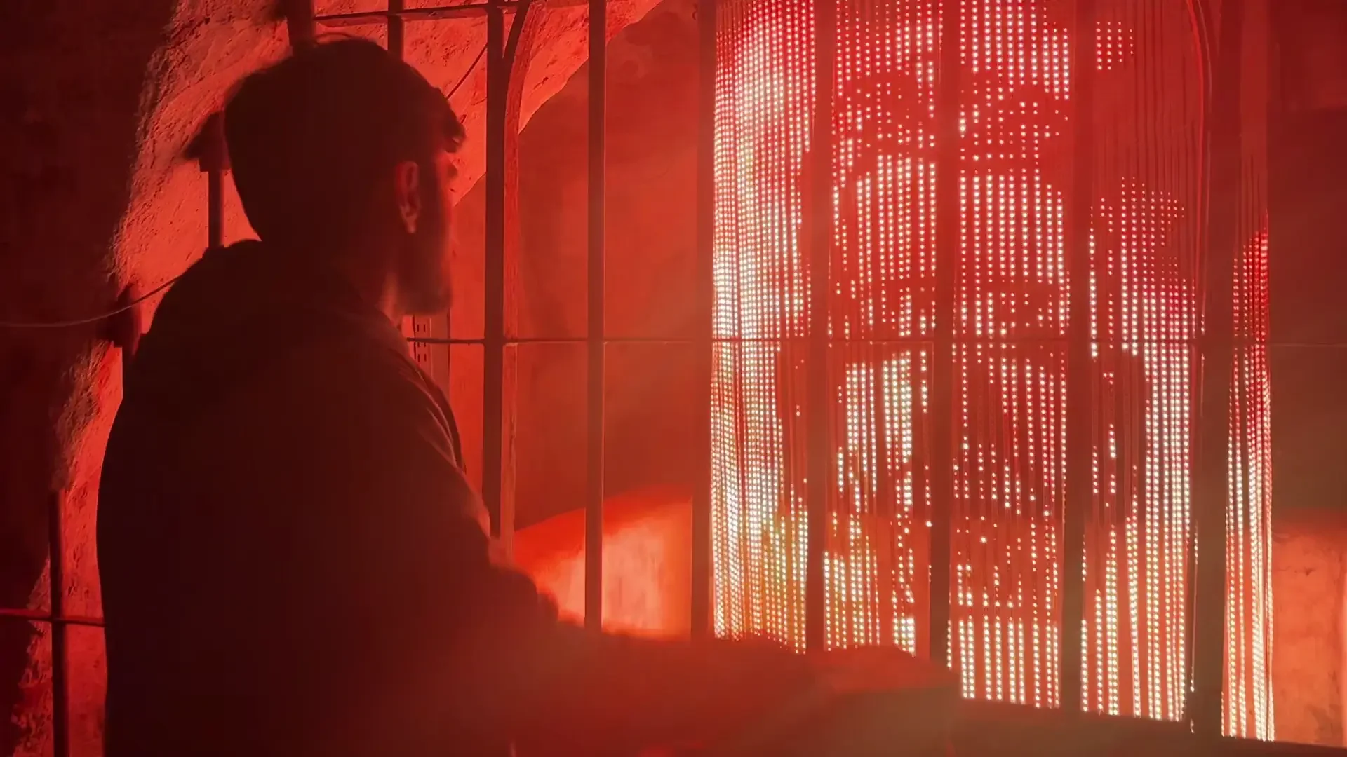 A person looking at a large cylindrical panel of red LED lights, viewed through a metal grid, in a dimly lit room with reddish walls.