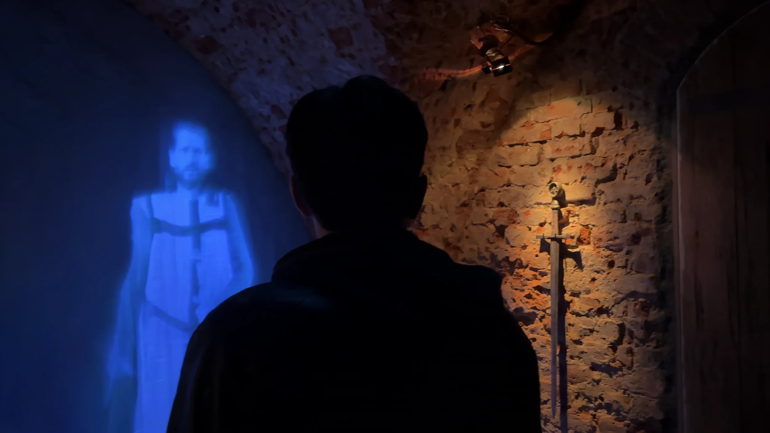 A person with short dark hair, viewed from behind, is looking at a bluish projection of Jesus Christ on a wall. The wall is made of rough bricks and there is a sword and cross mounted on the wall beside the projection.