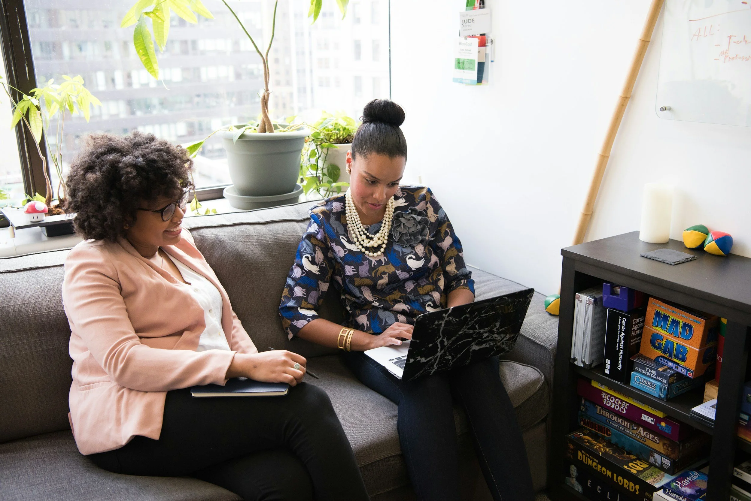 Two women sit on a gray couch in a modern office or lounge, looking at a laptop. The woman on the left has curly hair, glasses, and is wearing a peach blazer with a white shirt, taking notes on a tablet. The woman on the right has her hair in a bun, wearing a patterned dress with flower and animal designs, and a pearl necklace. Behind them is a window with a view of a cityscape and potted plants.