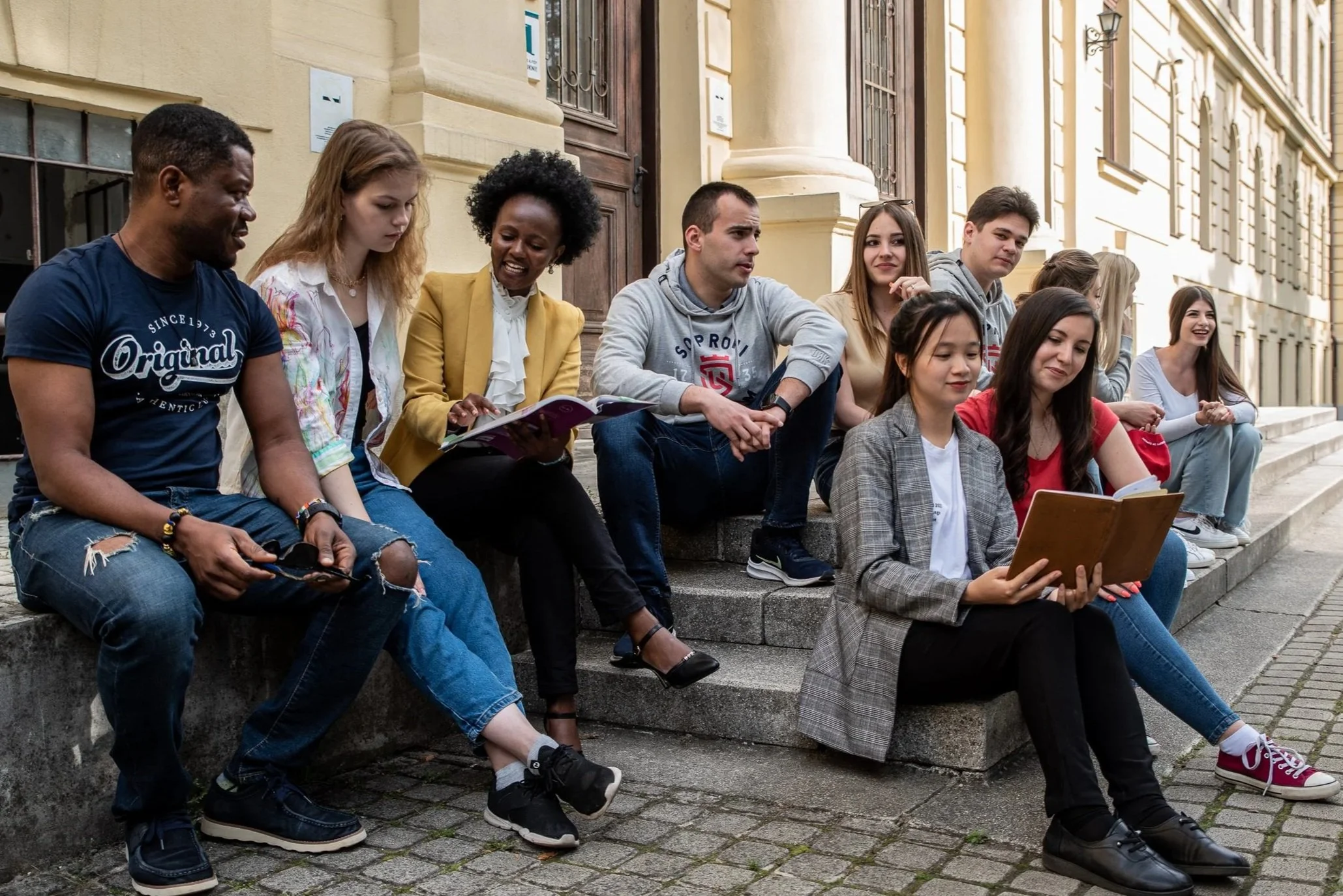Group of diverse young adults sitting on steps outside a building, some reading books, others talking or smiling.