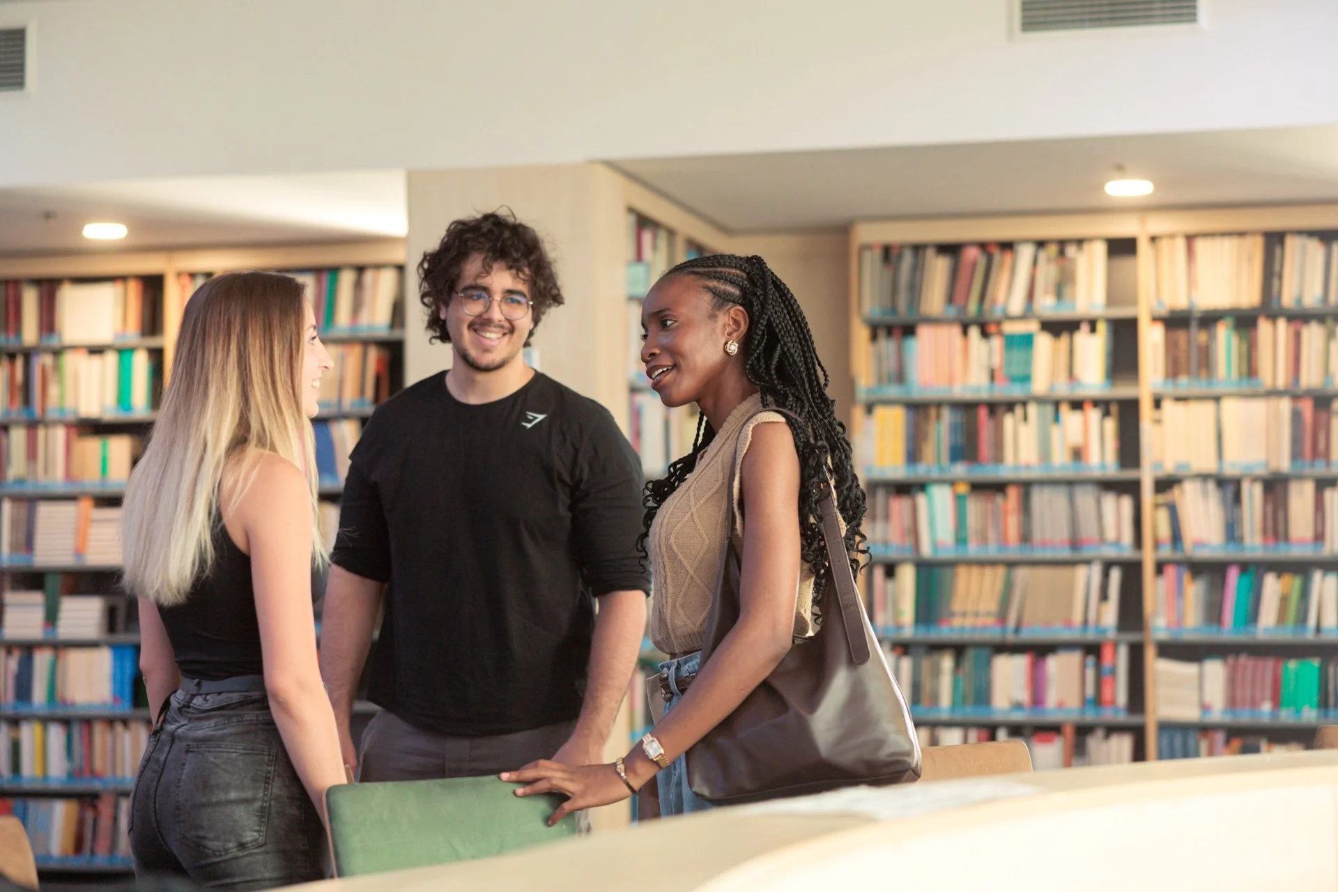 Three young adults talking and smiling in a library with bookshelves in the background.