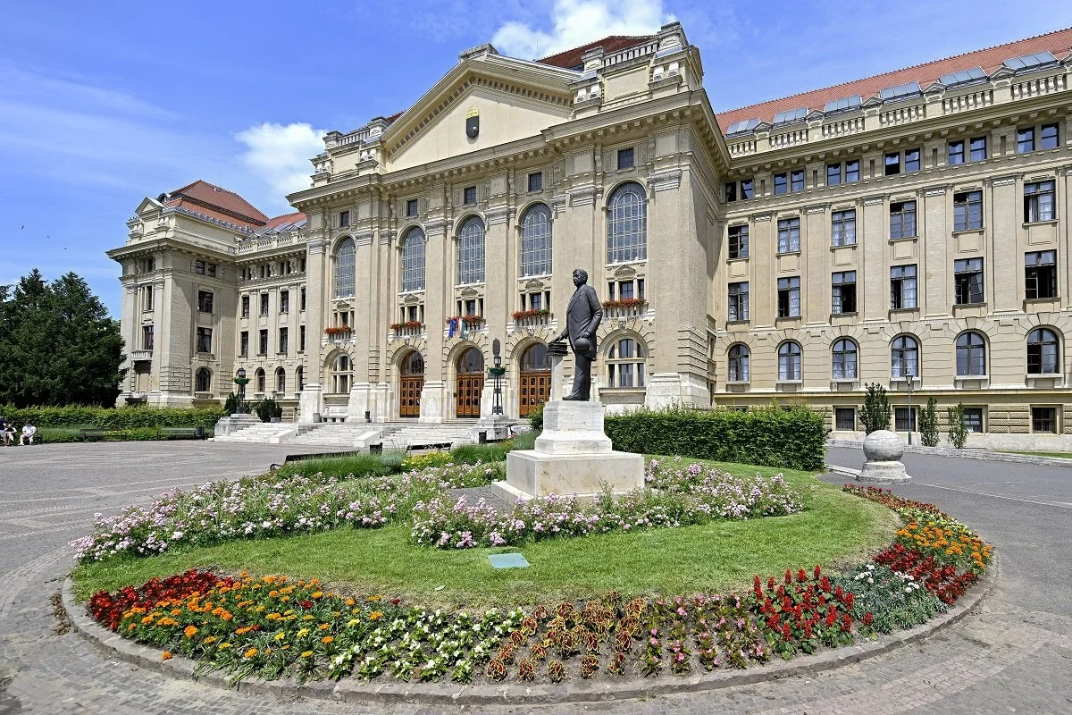A grand beige building with tall arched windows and a red roof, with a statue of a man holding a book in front surrounded by colorful flowers and greenery.