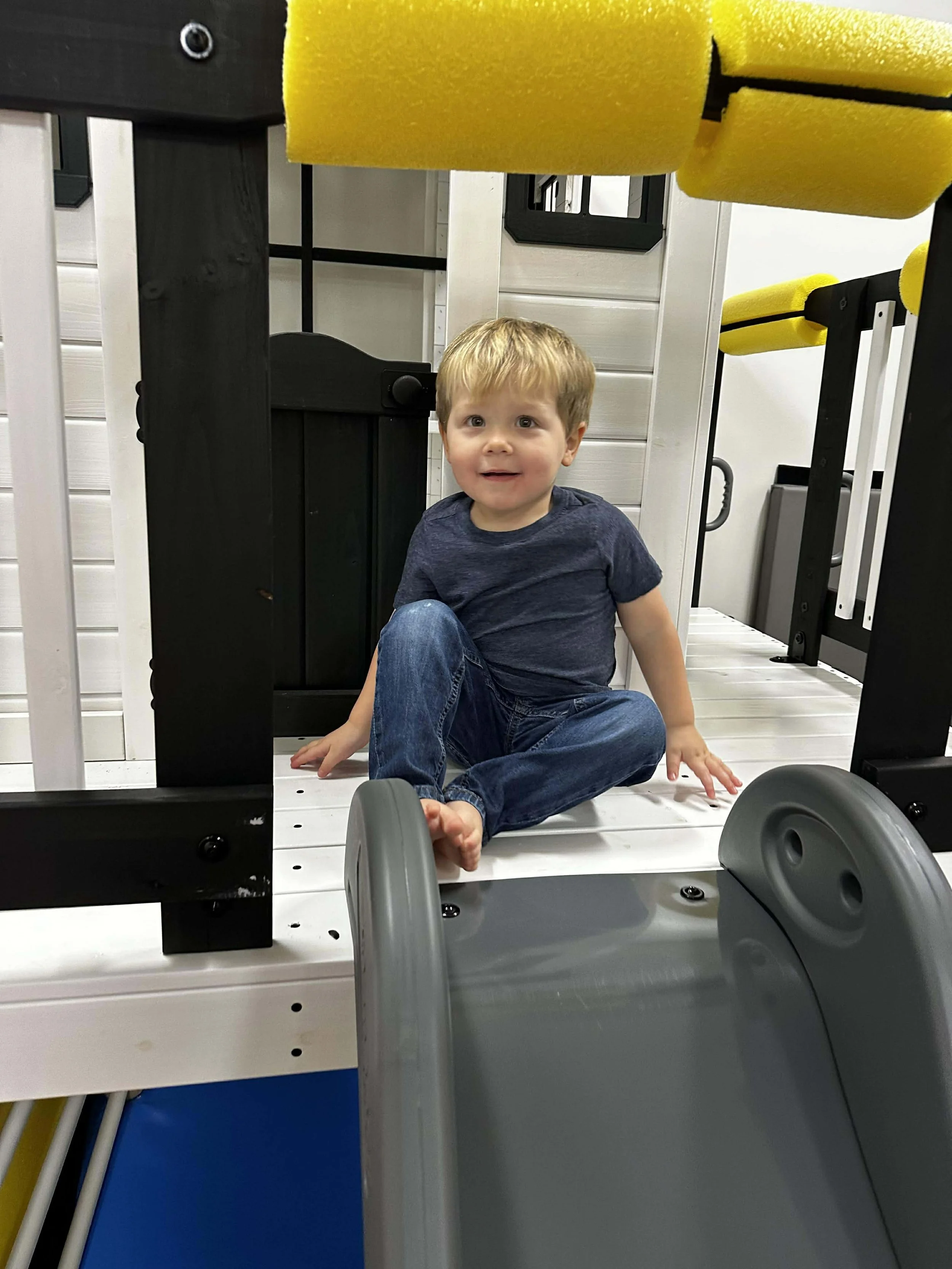 A young boy with blonde hair wearing a navy t-shirt and jeans sitting on a white play structure with a gray slide in an indoor playground.