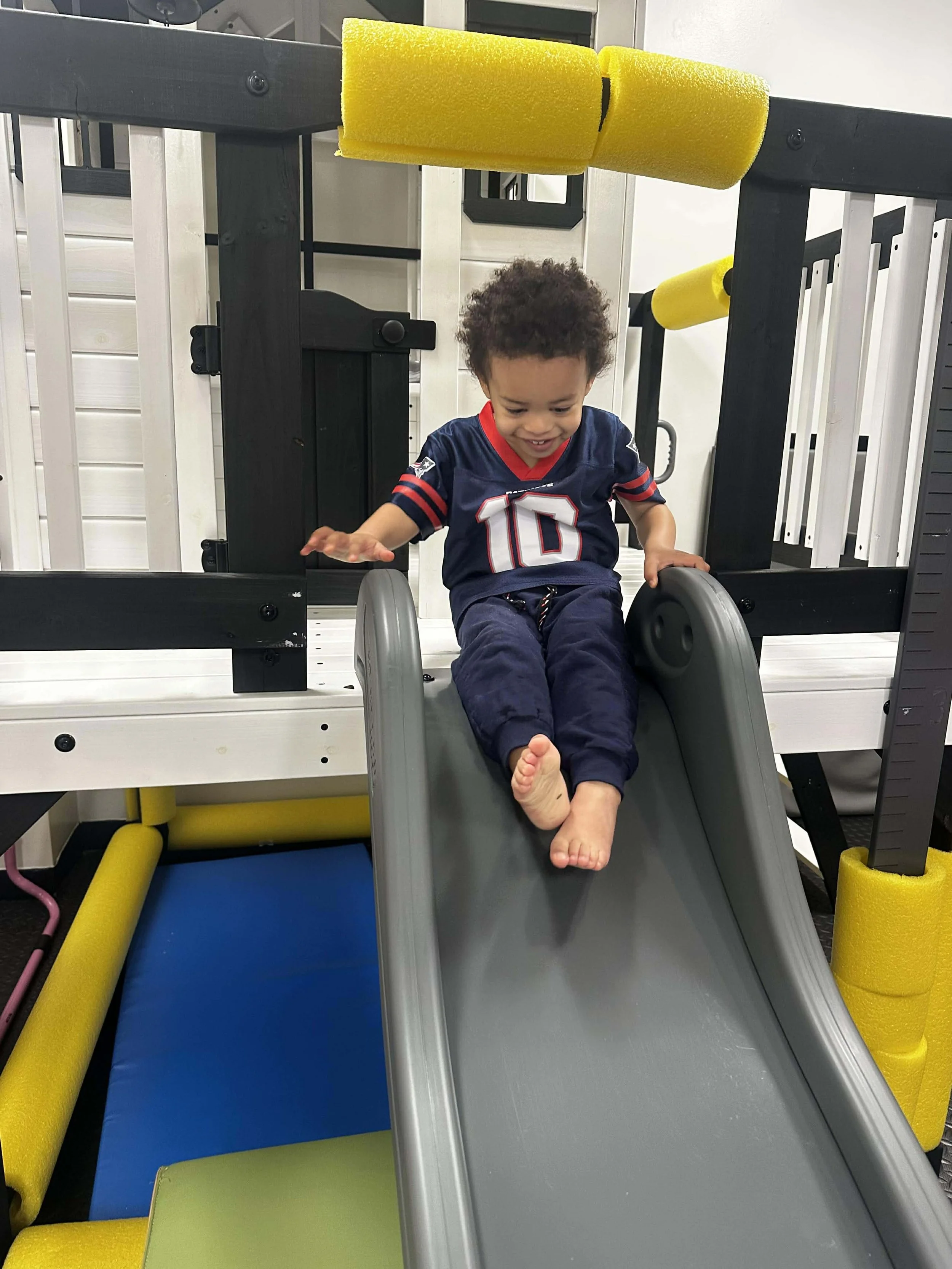 A young child with curly hair smiling as he slides down a small gray slide in an indoor play area, with colorful padded barriers around.