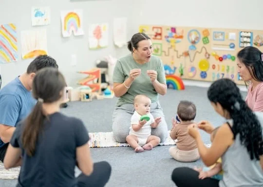 A group of women and babies sitting on the floor in a preschool classroom, engaging in a circle activity with a teacher leading.