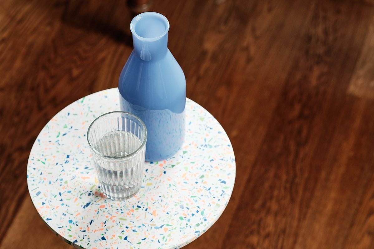 Eine blaue Flasche und ein Glas mit Wasser stehen auf einem rundem, buntem Tablett auf einem Holztisch. Versorgung mit Wasser in den Therapiestunden in der Psychotherapiepraxis Dialogerie in Wien 1050.