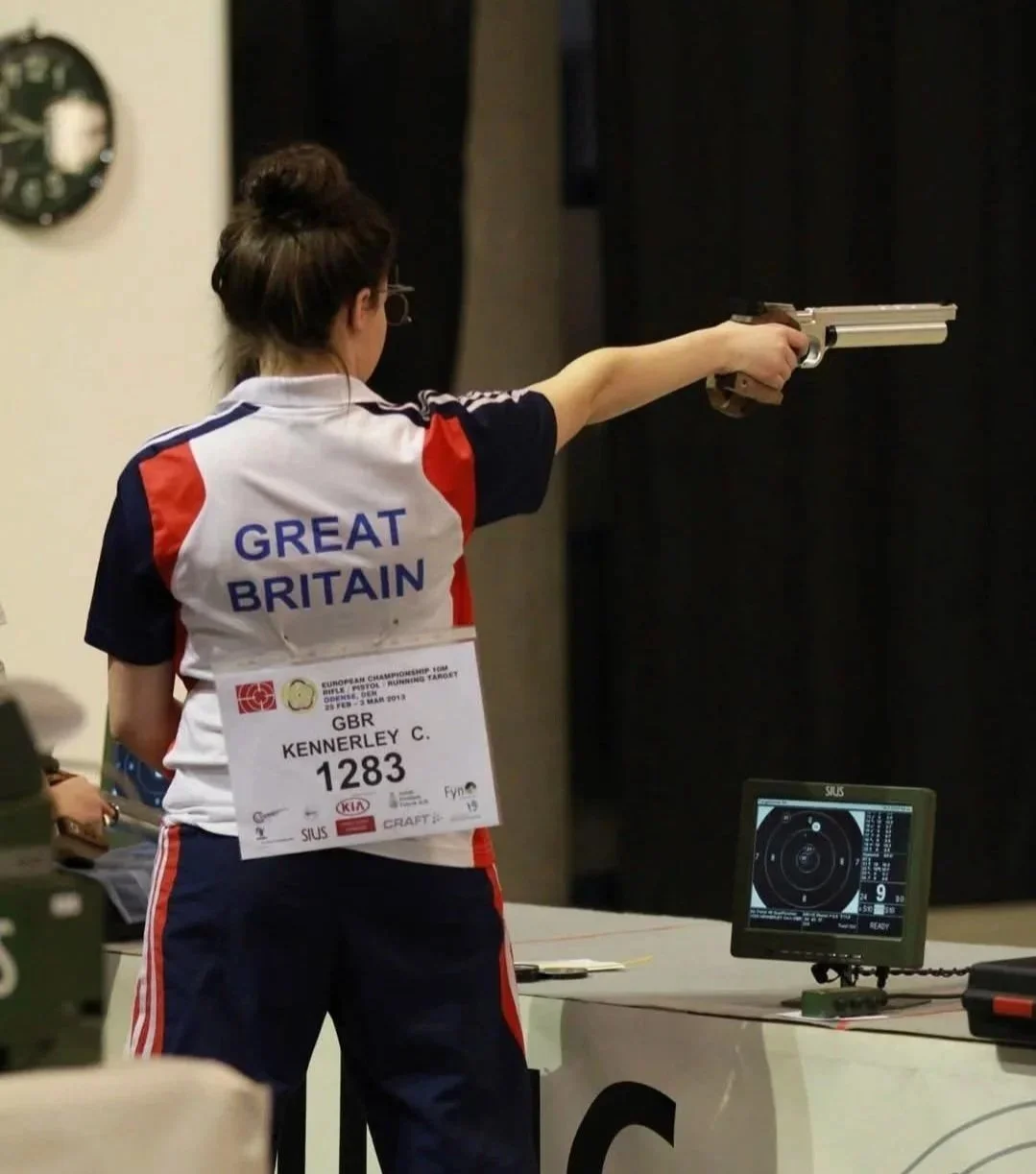 A woman wearing a Great Britain sports uniform is shooting a pistol at a shooting range, with her right arm extended and aiming at a target.