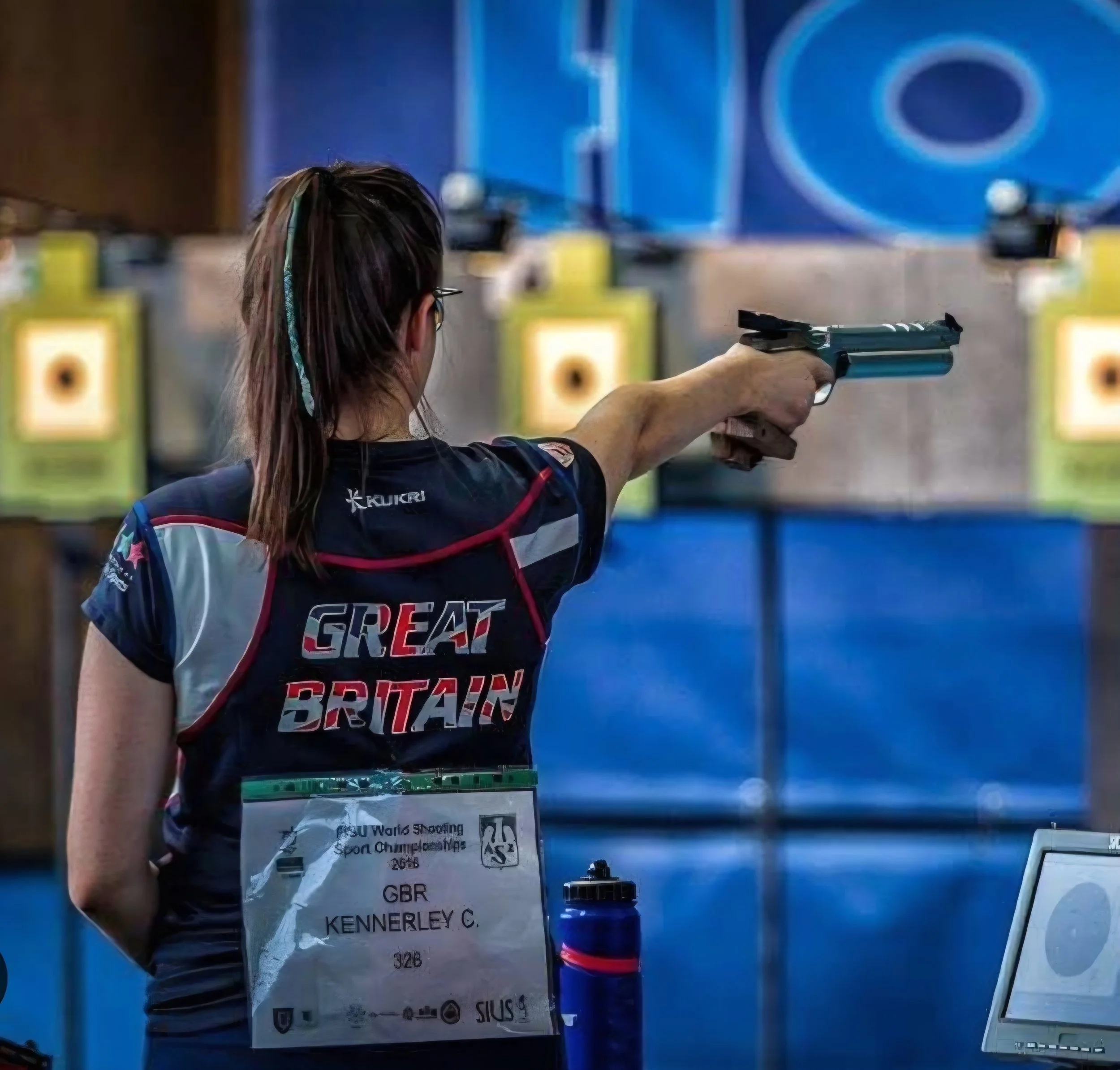 A young woman with brown hair wearing glasses in a Great Britain team shirt, shooting a handgun at a target in an indoor shooting range. She has a name tag with her details attached to her shirt.