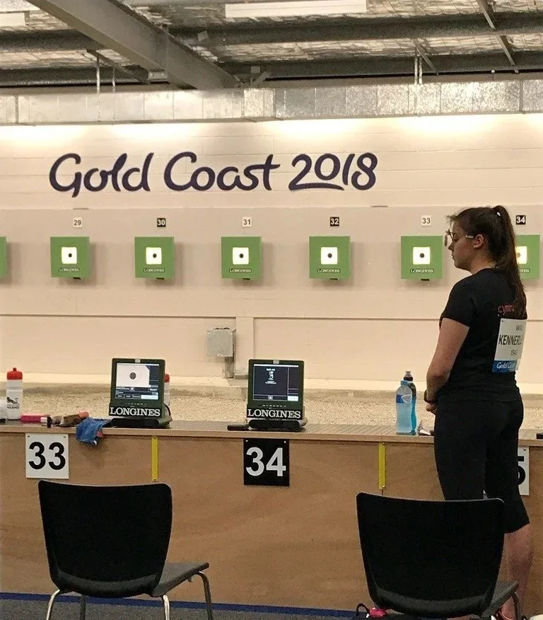 Indoor shooting range with targets labeled 29 to 34, set for the Gold Coast 2018 event, with two computers and shooting gear on the table, and a woman standing at position 34 in athletic attire.