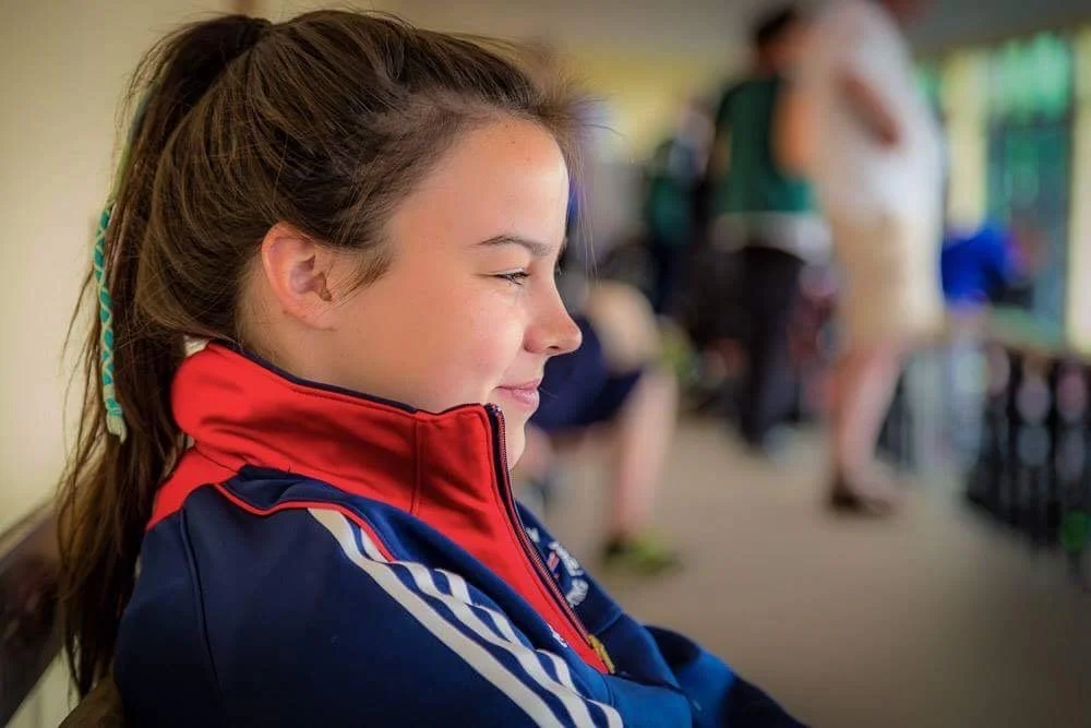 A young girl with a ponytail smiling while sitting indoors, wearing a red and blue sports jacket.