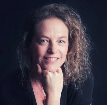 Photo de Laurence Leduc, avec des cheveux bouclés, souriant et posant avec sa main sous son menton, contre un fond sombre.