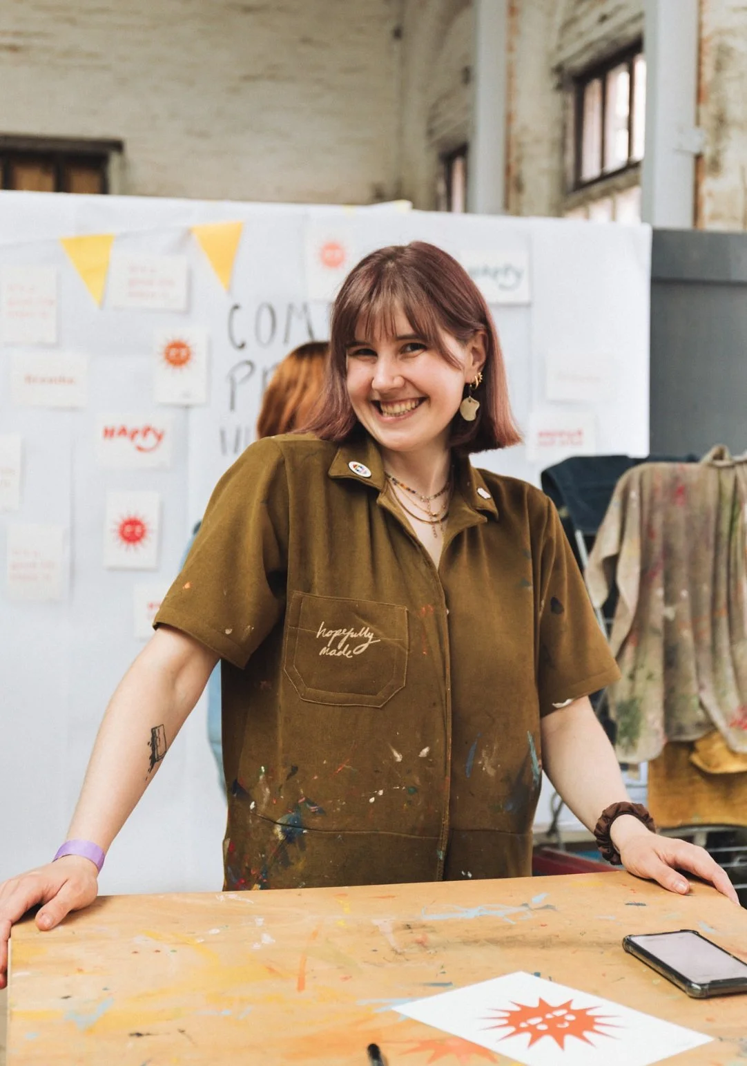 A smiling woman with short brown hair and earrings standing behind a table in an art studio. She is wearing a paint-splattered brown jumpsuit The background features hand printed artworks on a white display board.