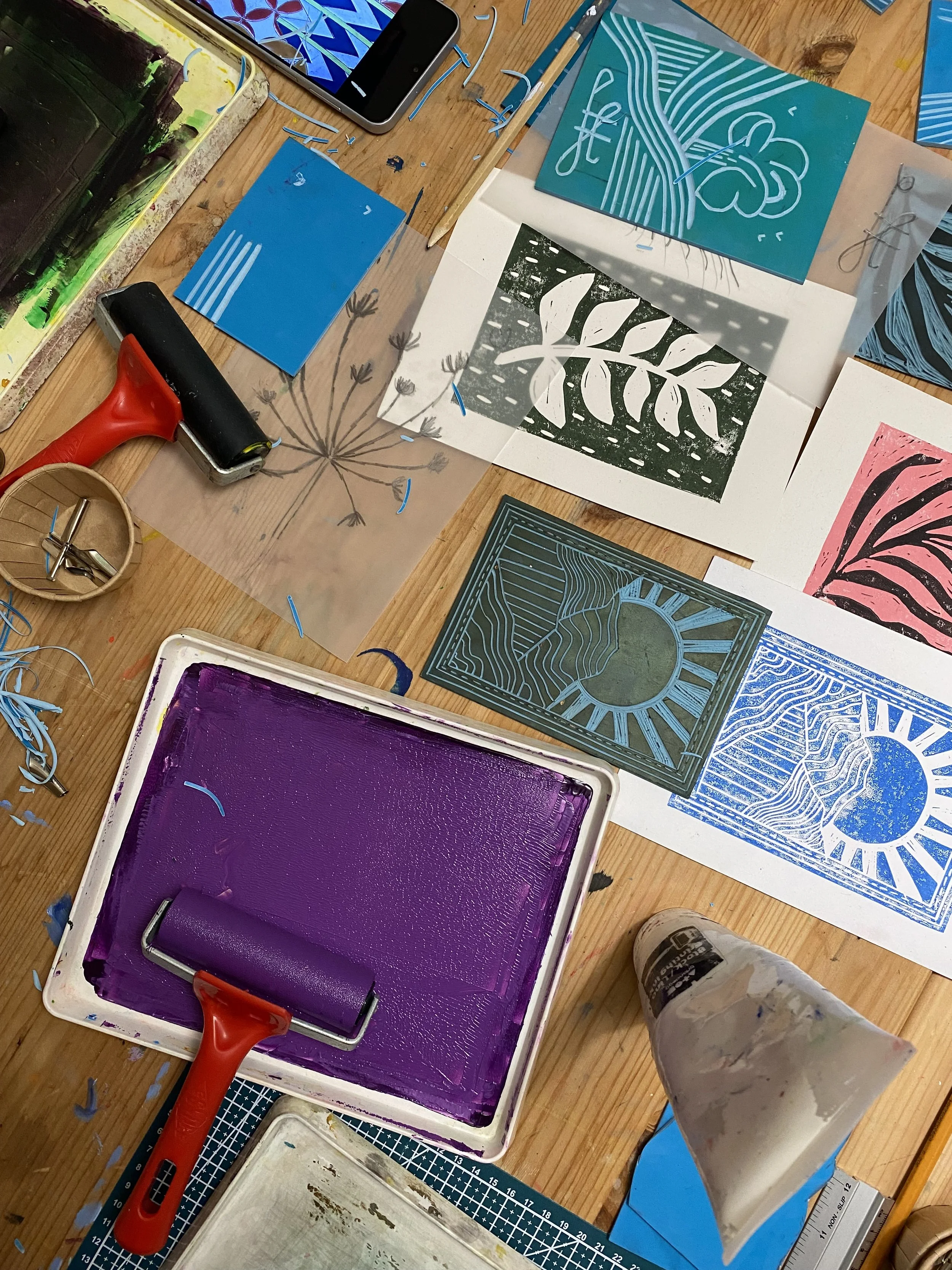 An art workspace with various stencils, paint, and tools spread on a wooden table, including stencils of abstract patterns and nature motifs, a small roller, various paint colours, a smartphone, and Lino printing supplies.